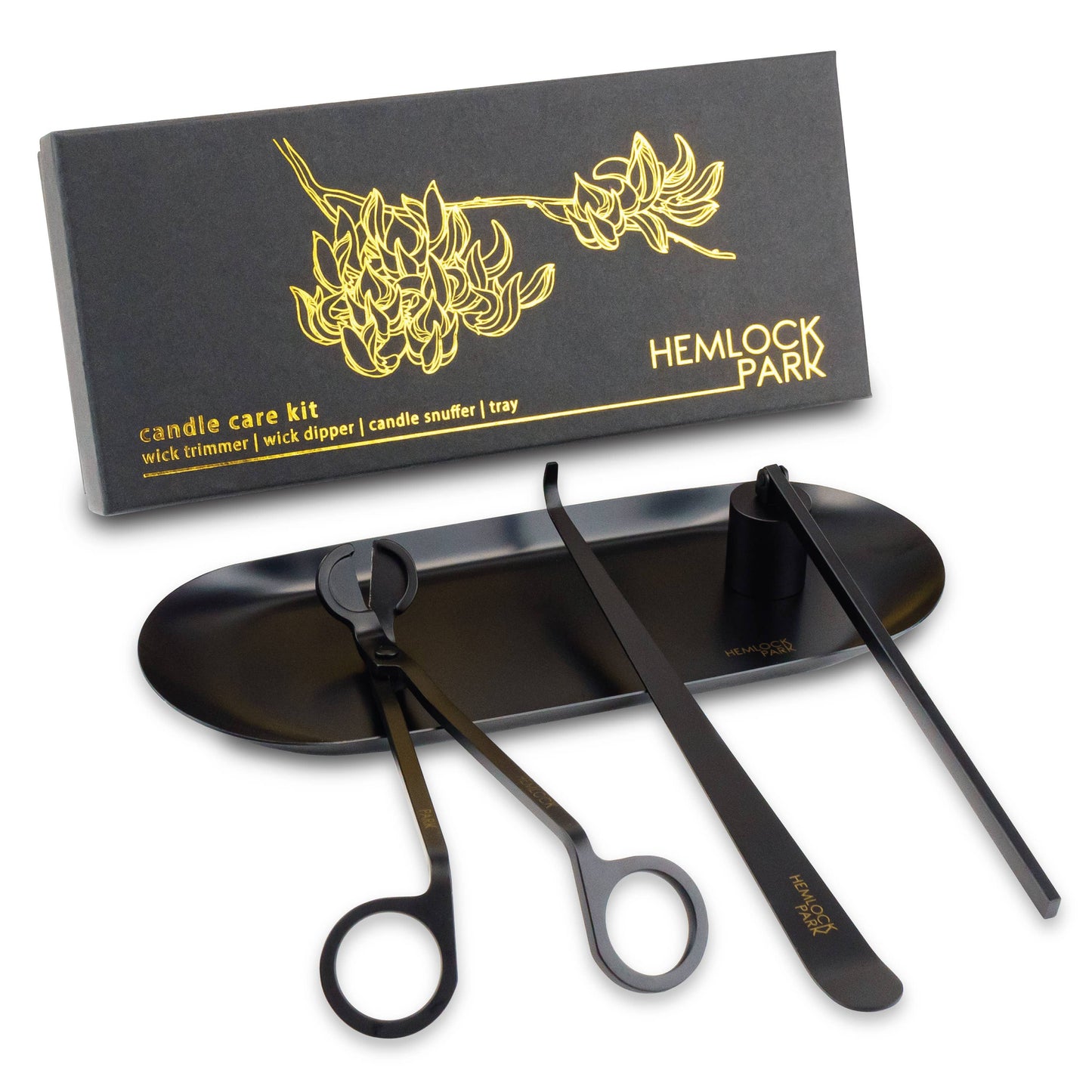 A black Candle Care Kit with a gold floral design on the box includes a candle wick trimmer, dipper, snuffer, and tray—all neatly arranged before the elegant packaging.