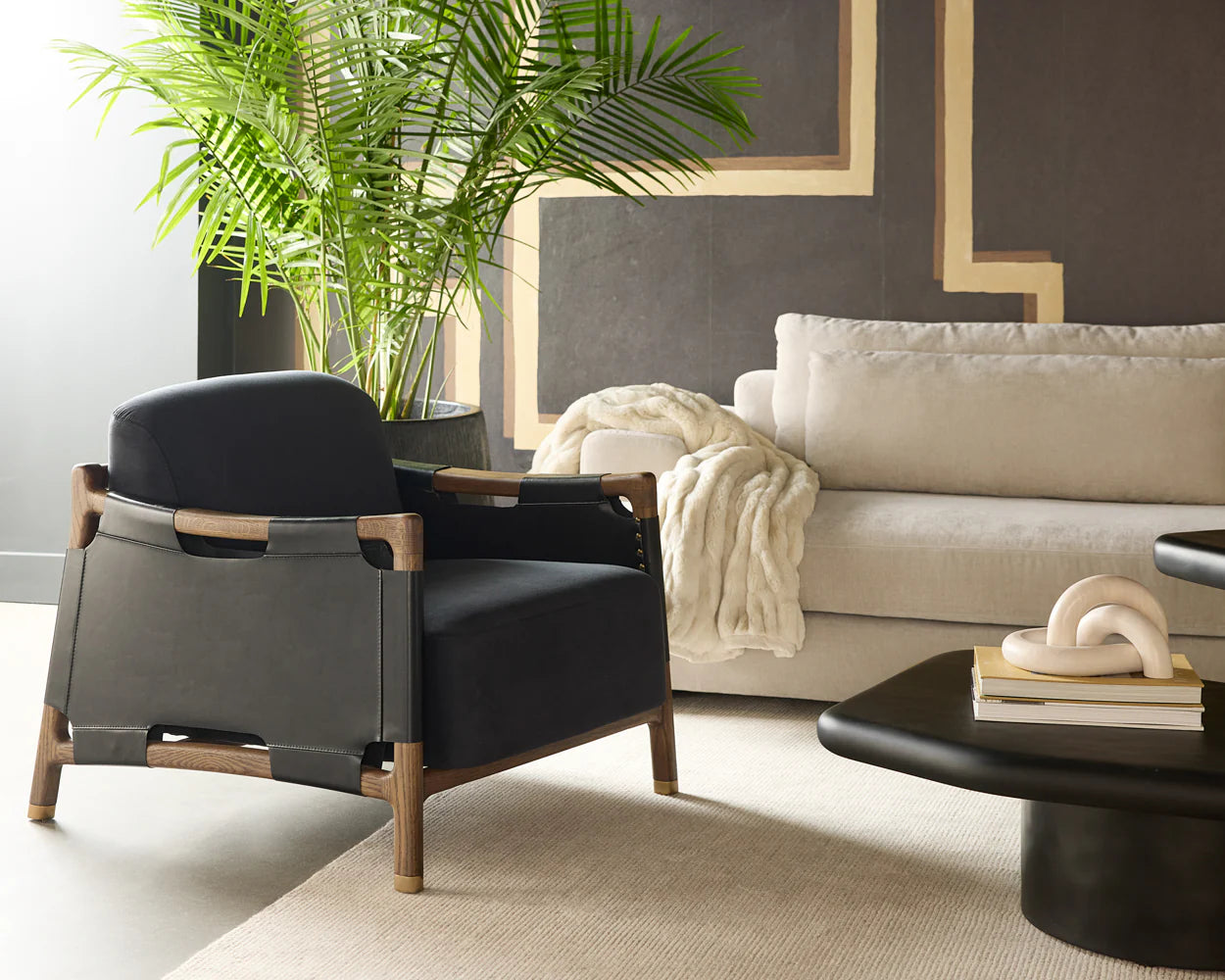 A modern living room with the Black Leather Lounge Chair featuring a solid oak frame and Italian leather straps, a light beige sofa with a cream blanket, a black coffee table with books and sculpture, and a tall green plant in the background.