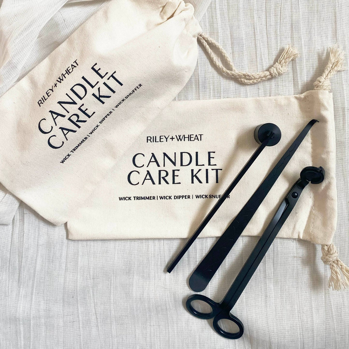 The Candle Care Kit includes a sleek black wick trimmer, dipper, and snuffer on a white cloth, alongside two cream Riley + Wheat Candle Care Kit pouches.