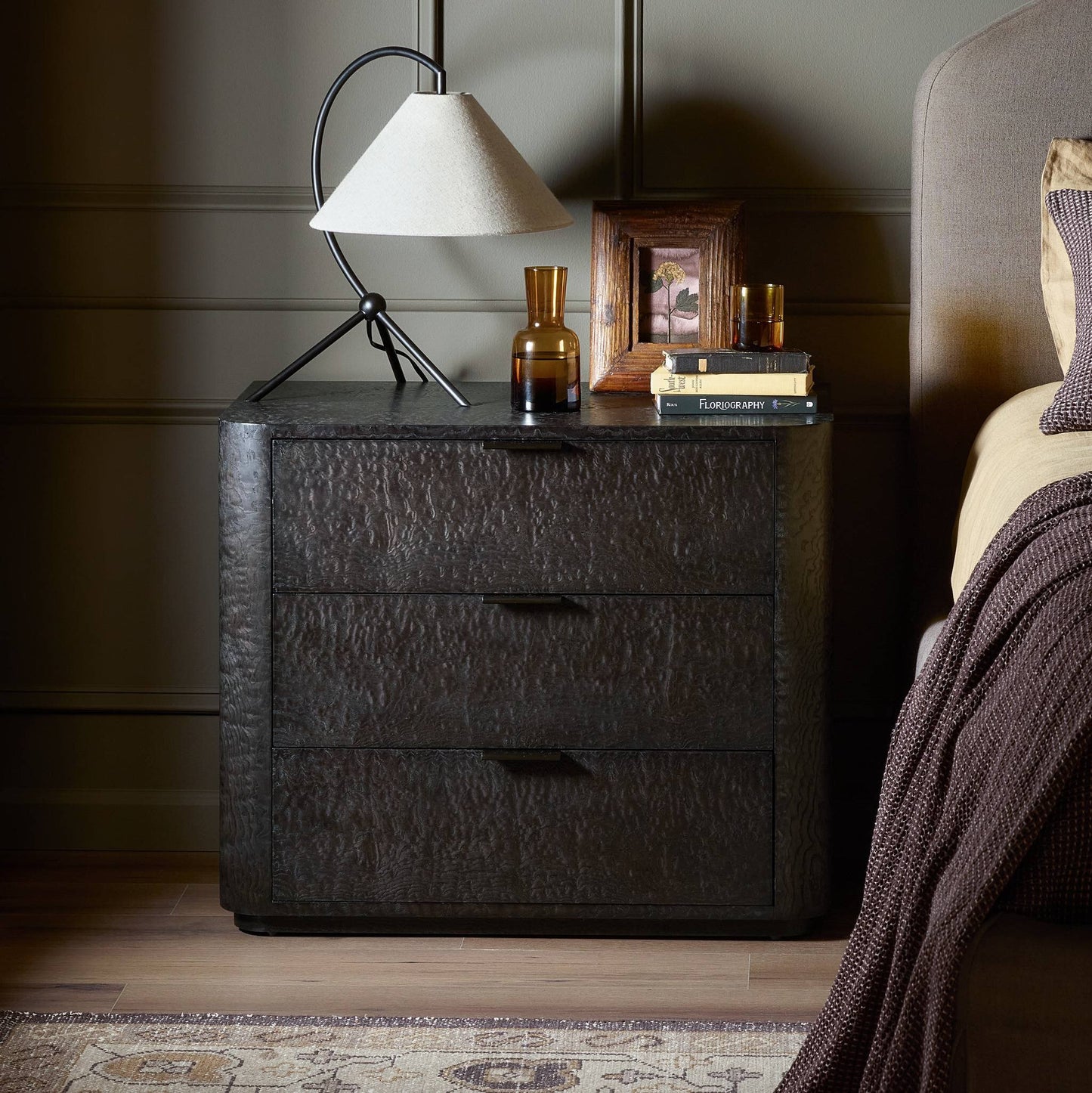 The Stenson 3 Drawer Nightstand (32.00"w x 20.00"d x 26.50"h) holds a modern lamp, two amber glass vases, a framed photo, and books beside a bed with brown bedding in a cozy, softly-lit bedroom.