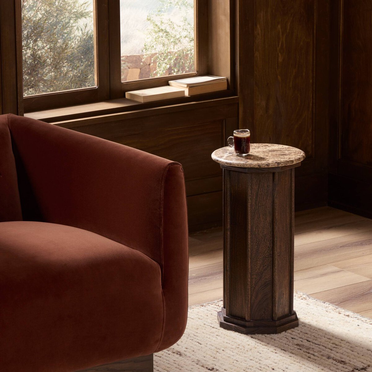 A cozy living room with a brown velvet sofa and the Pavlo End Table (11.00"w x 11.00"d x 21.00"h) featuring a marble top holding coffee, wooden paneled walls, and sunlight streaming onto books along the window sill.
