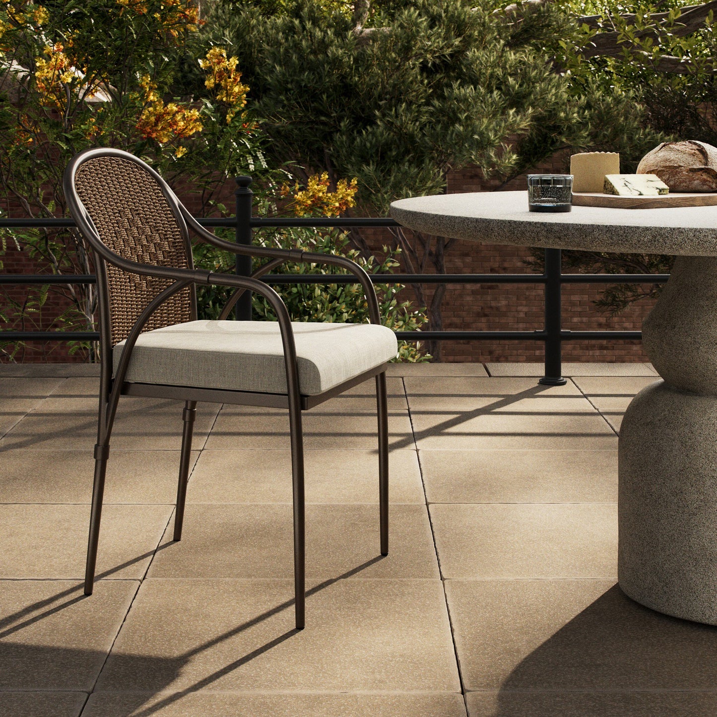 The Graciela Outdoor Dining Chair, featuring a woven back and beige cushion (21"w x 22.5"d x 35"h), sits beside a round stone table with bread, cheese, and water on a tiled terrace surrounded by greenery.