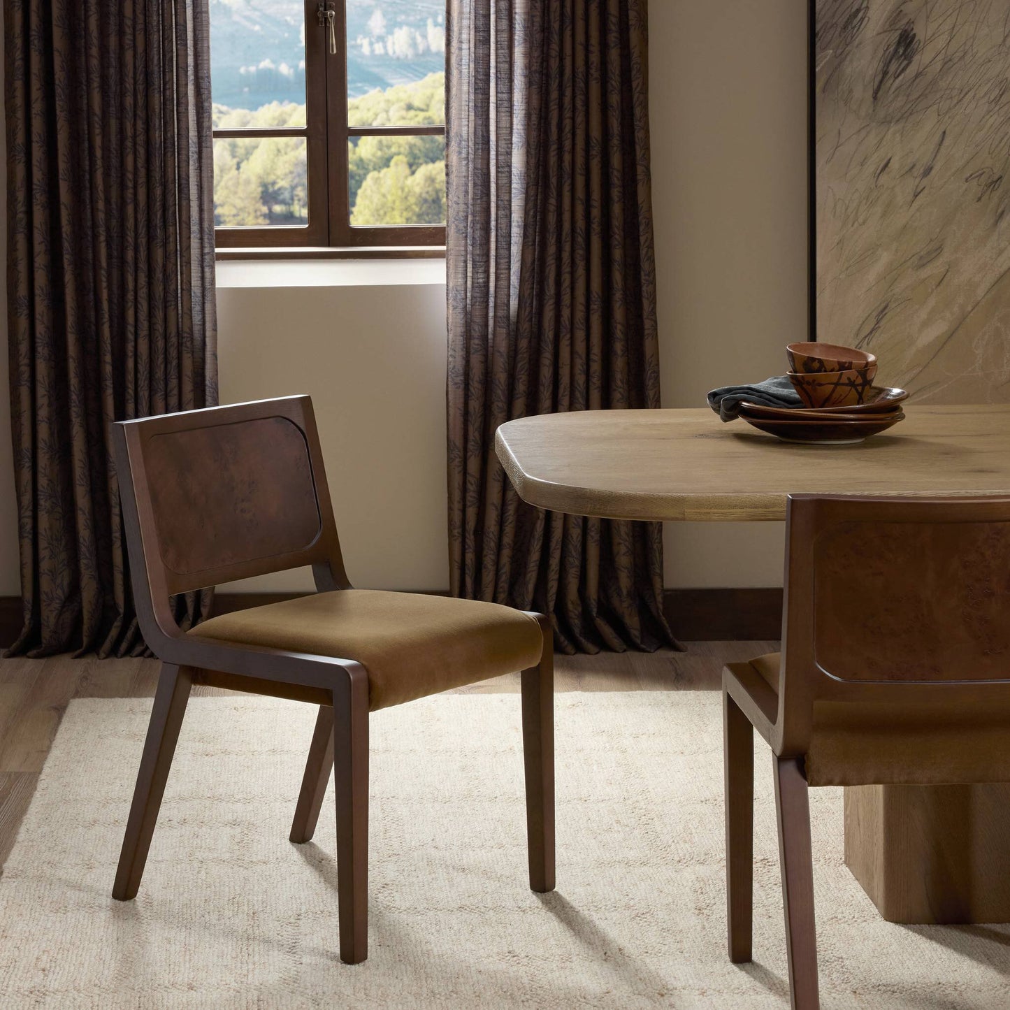 A modern dining room featuring a wooden table, two Corvo Dining Chairs in brown, a beige rug, dark window curtains, and ceramic bowls and plates decorating the table. Trees are visible through the window.