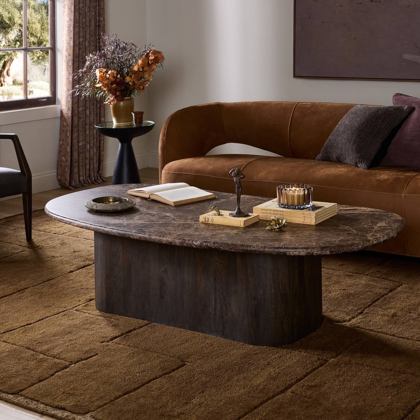 A modern living room features a 65-inch brown velvet sofa, the Tori Coffee Table with books and decor, a brown rug, and a side table with a vase of dried flowers near the window.