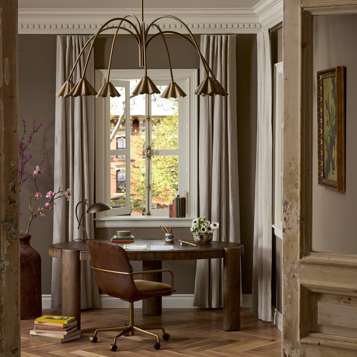 A cozy home office has a round wooden desk, brown swivel chair, and the modern Amabella Chandelier in Aged Antique Brass. A table lamp, flowers, and books by the window with beige curtains complement warm brown walls and herringbone floor.