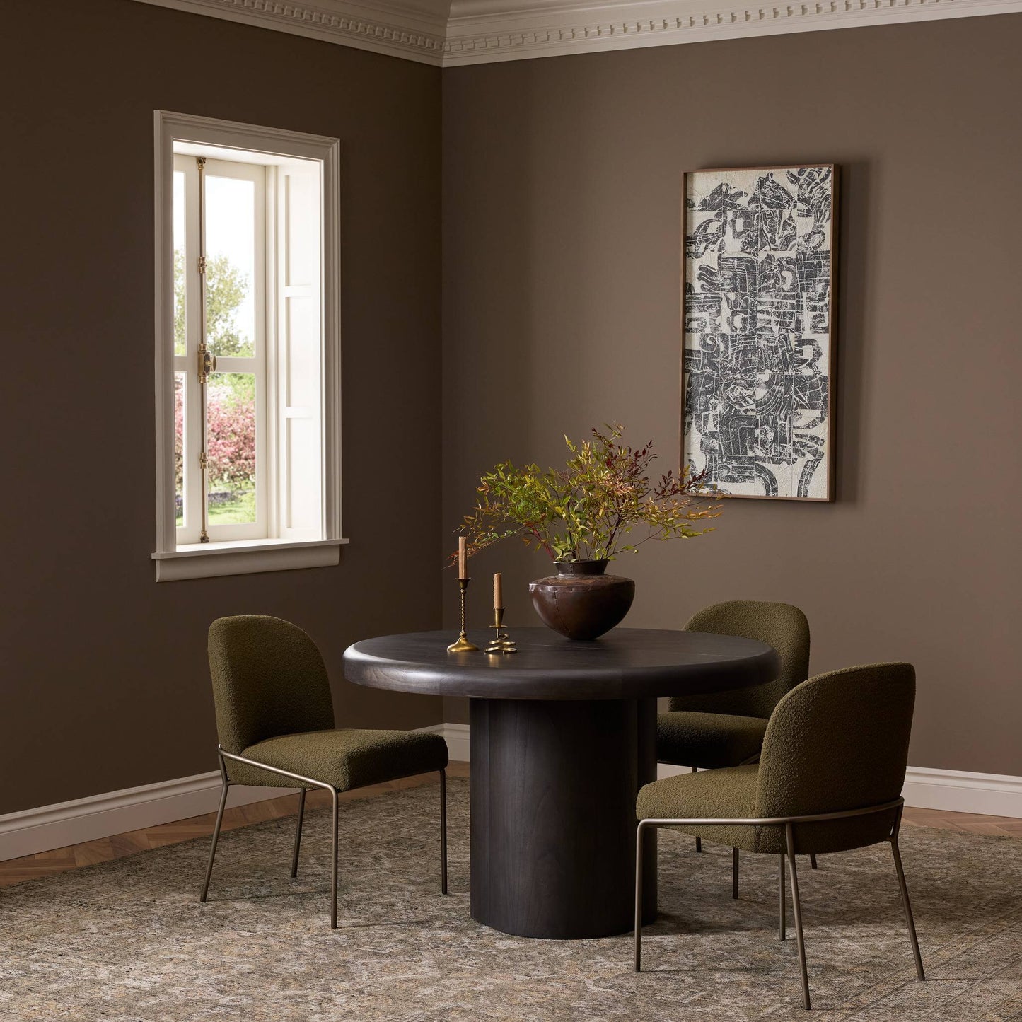 The Pemble Extension Dining Table (48x48) with two green cushioned chairs sits in a brown-walled room, decorated with a vase of branches, candlesticks, framed art, and bright natural light streaming through the window.