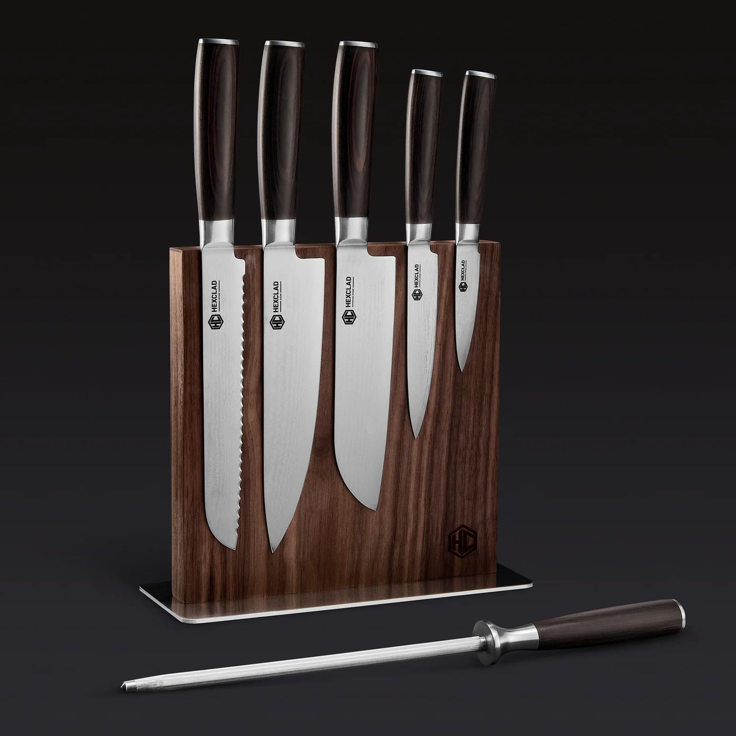 The Hexclad Steel Knife Block features five kitchen knives with dark handles displayed upright in a wooden magnetic block, plus a metal honing rod in front, all set against a dark background.