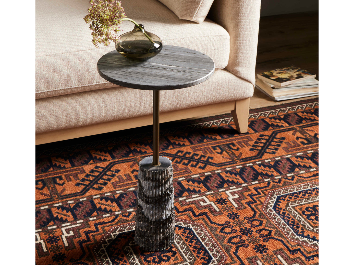 The Raven End Table, featuring a textured round base, stands on a patterned rug beside a beige sofa. This compact accent table is perfectly sized to hold a green glass vase with dried flowers and has space for a stack of books nearby.