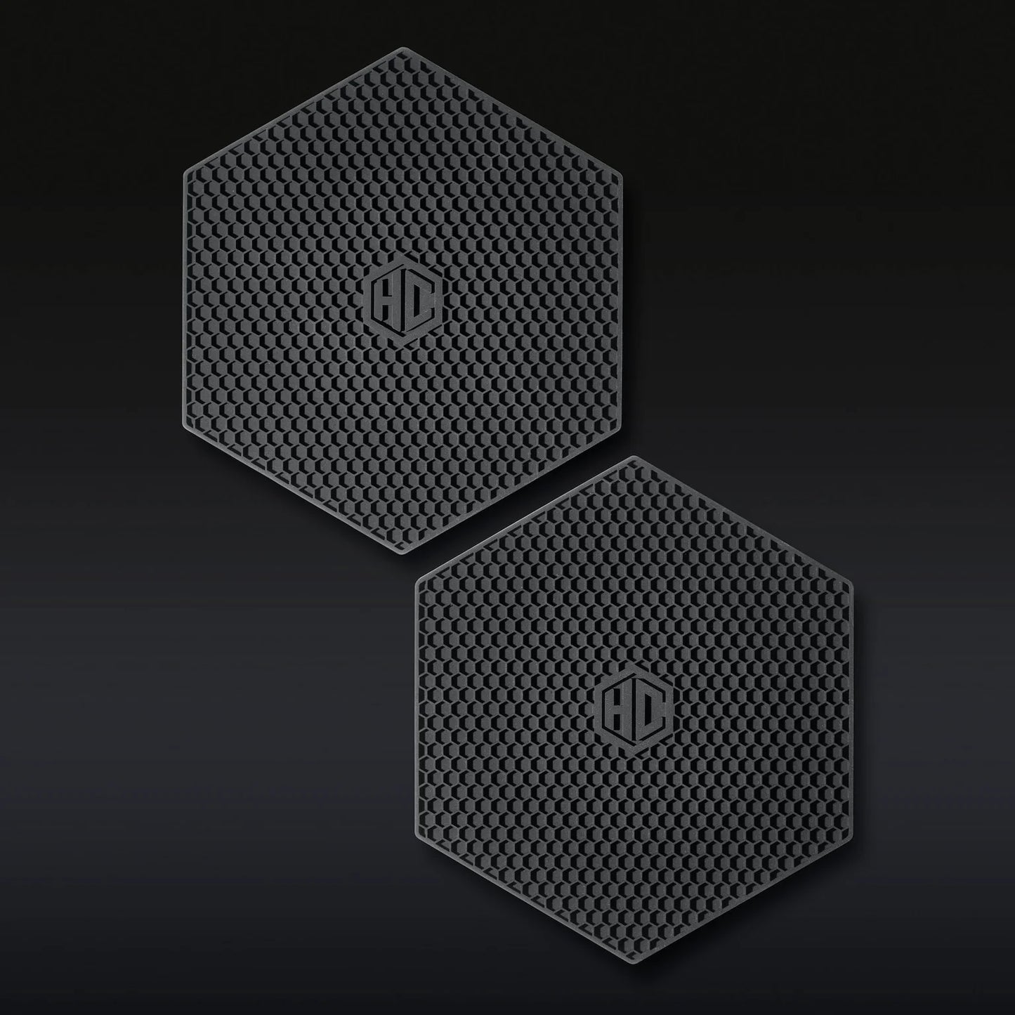 The Hexclad Trivet Set of 2 features black, hexagonal silicone pads with a honeycomb texture and a raised HD logo in the center, shown side by side on a dark background.