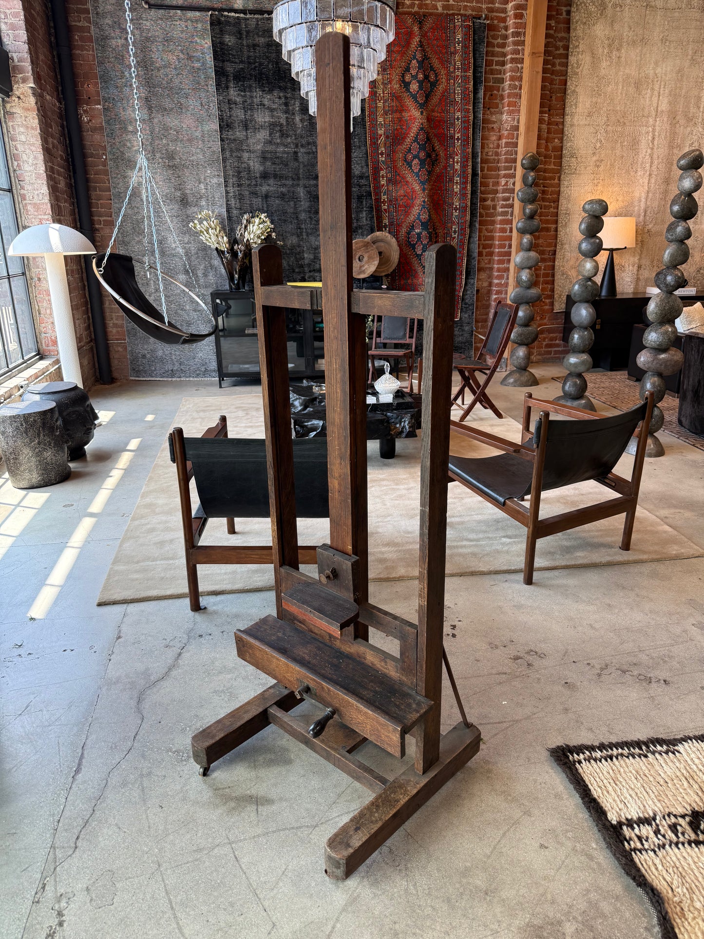 The 1800s Wood Art Easel adds charm to a stylish loft with brick walls, modern furniture, a hanging chair, decorative sculptures, and patterned rugs, all bathed in sunlight from large windows.