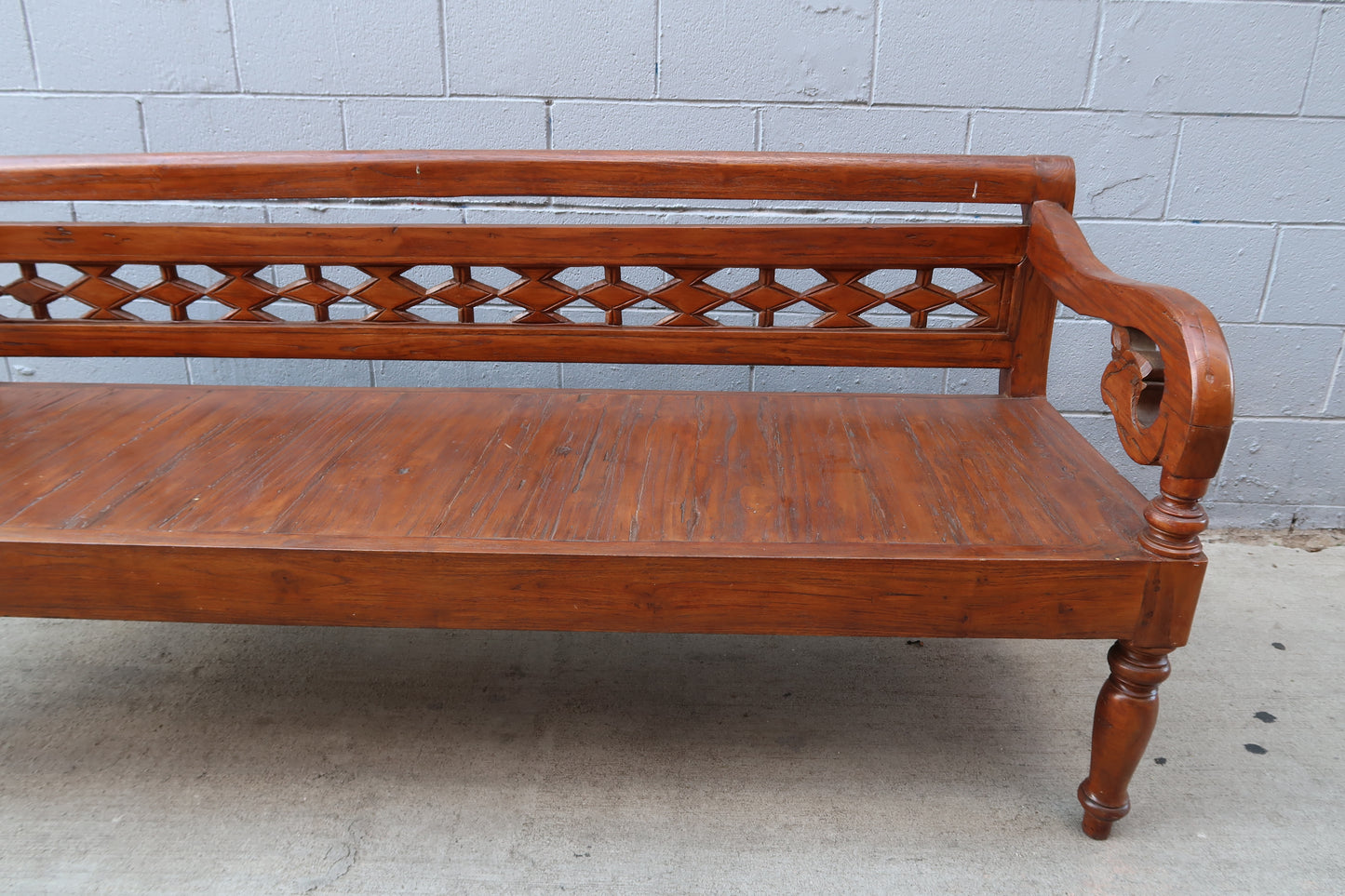 The Vintage Wood Carved Bench, ideal as accent furniture, showcases a diamond-patterned backrest and a right-side round armrest, placed on concrete against a light gray brick wall.