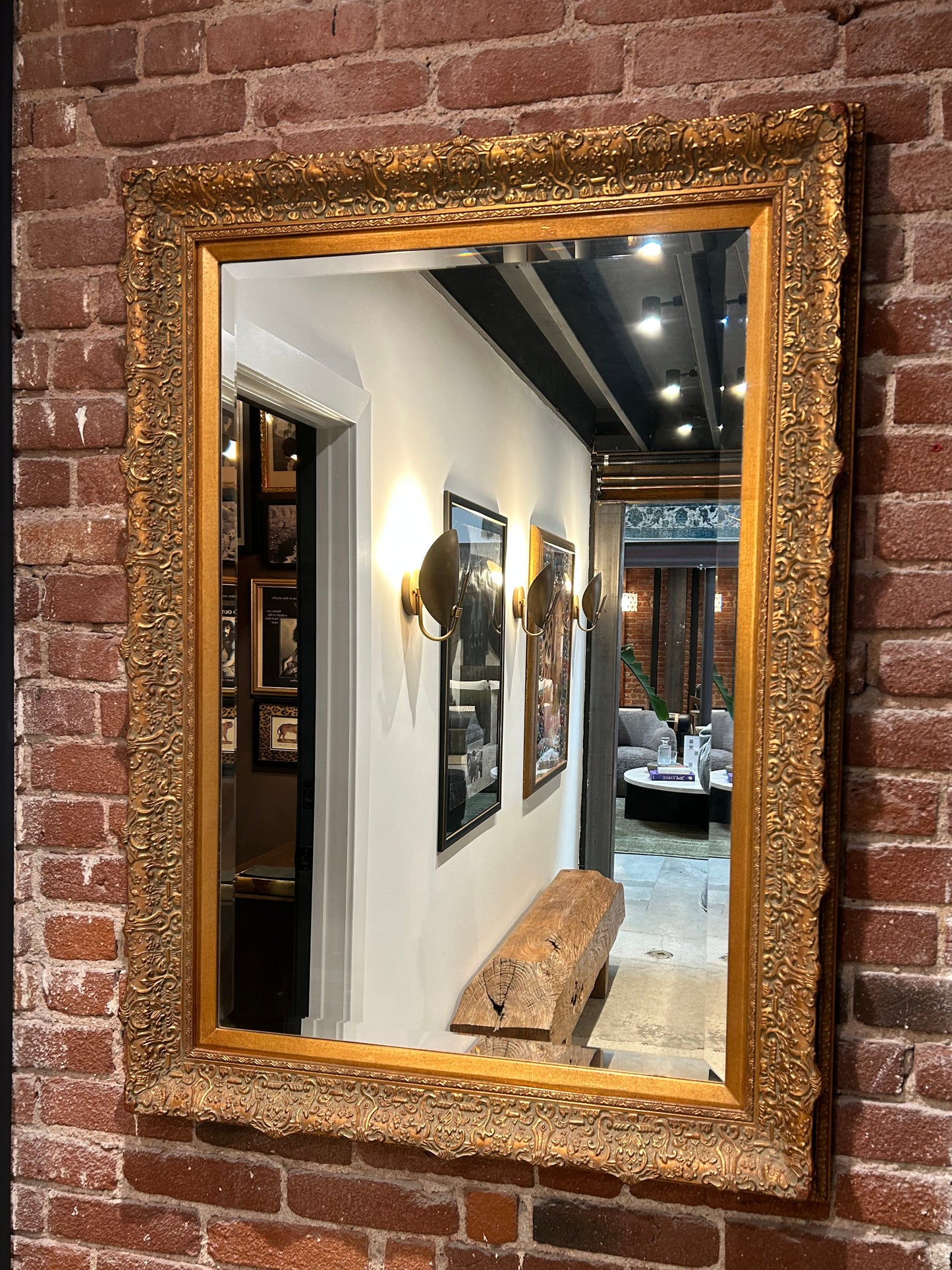 The Vintage Beveled Gilt Frame Mirror is mounted on a brick wall, reflecting a hallway with framed art, modern sconces, a wooden bench, and part of a cozy seating area.