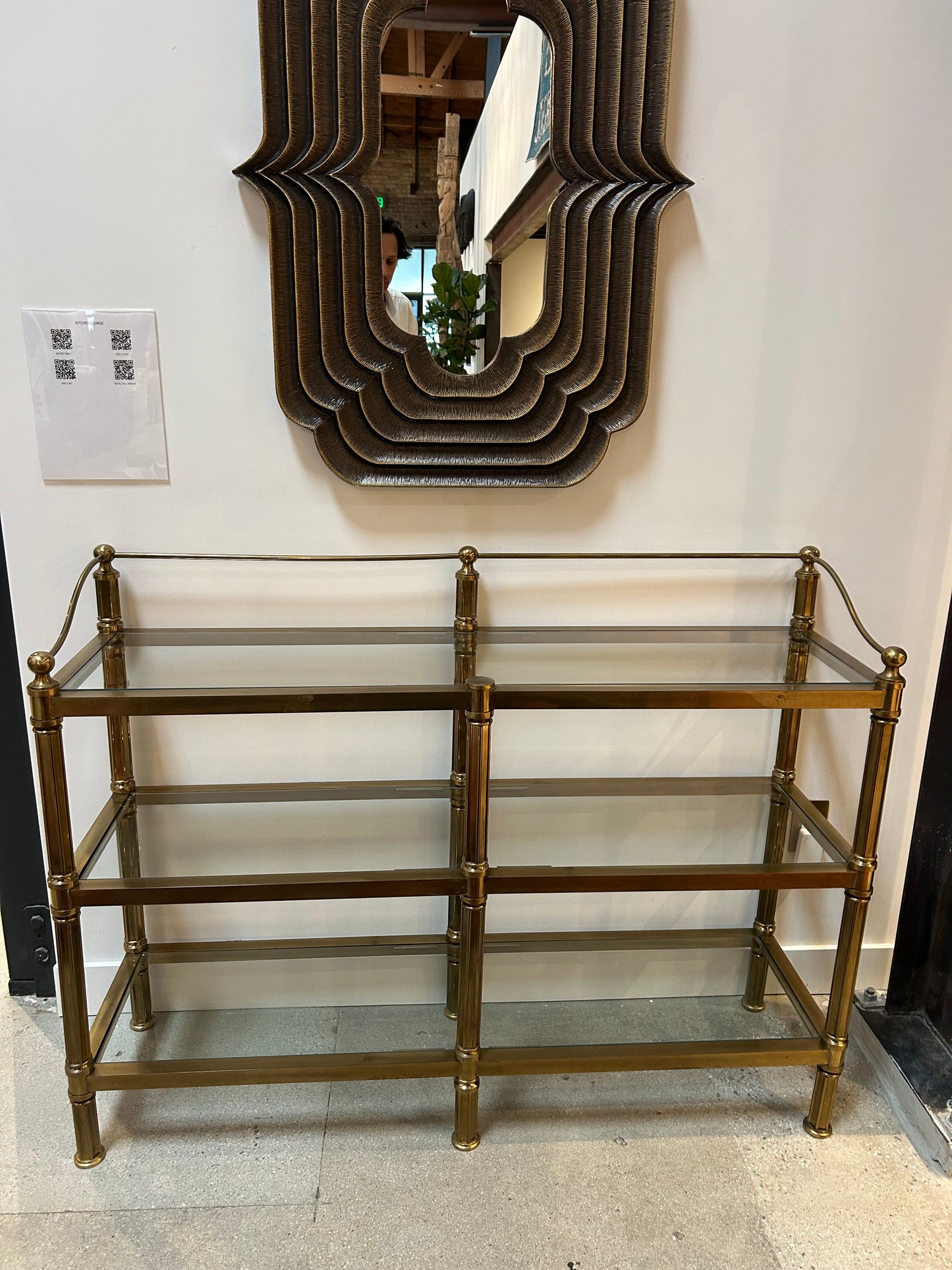 The Vintage Brass & Glass Console, measuring 52"W x 15"D x 39"H with three tiers, sits beneath a geometric layered wall mirror. Both are displayed against a white wall on a light-colored floor.
