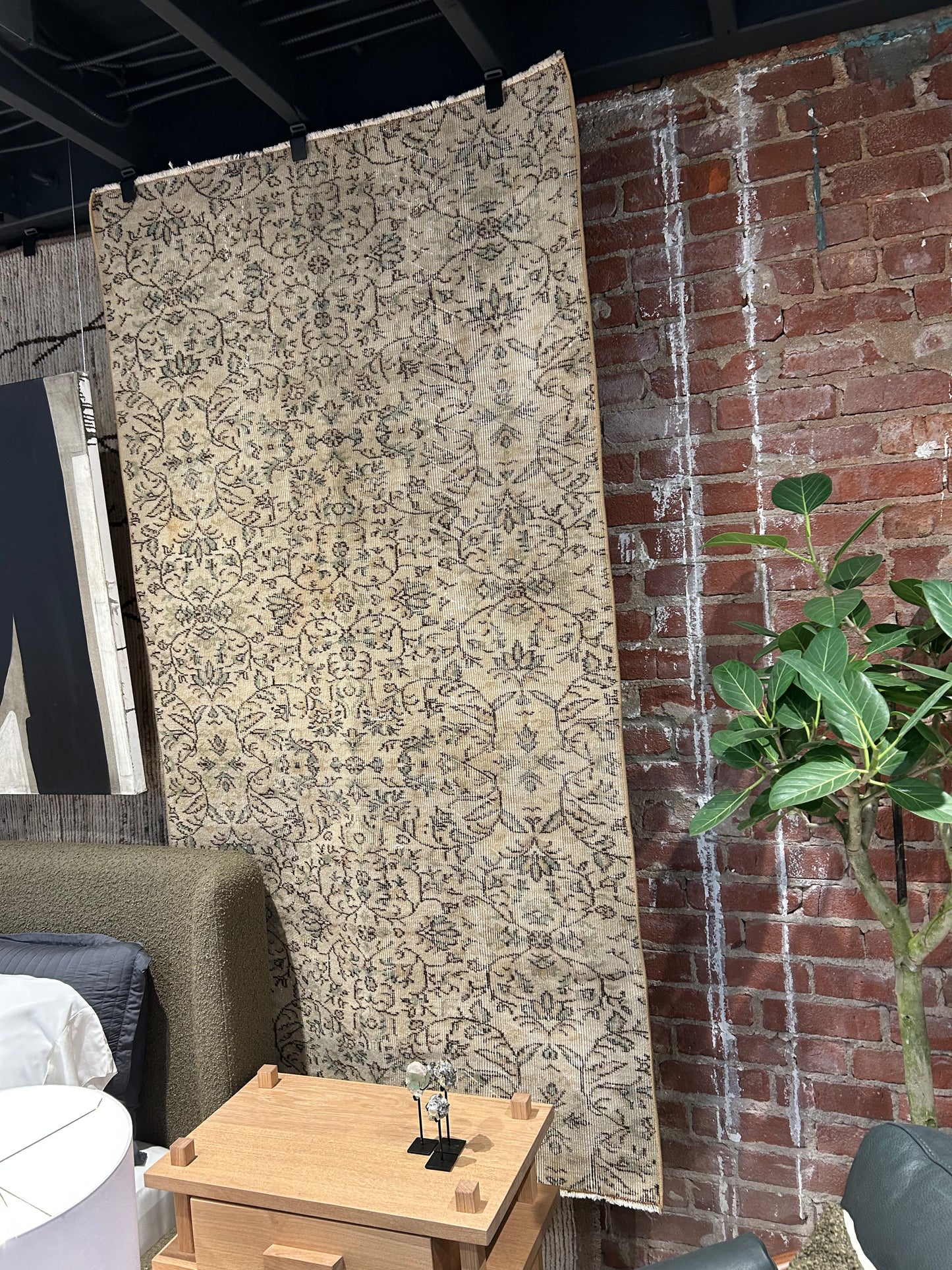 A Vintage Floral Runner Rug in tan and green with ornate patterns is displayed against an exposed red brick wall in a modern interior, next to a plant, wooden side table, and near an 80-inch-tall door.
