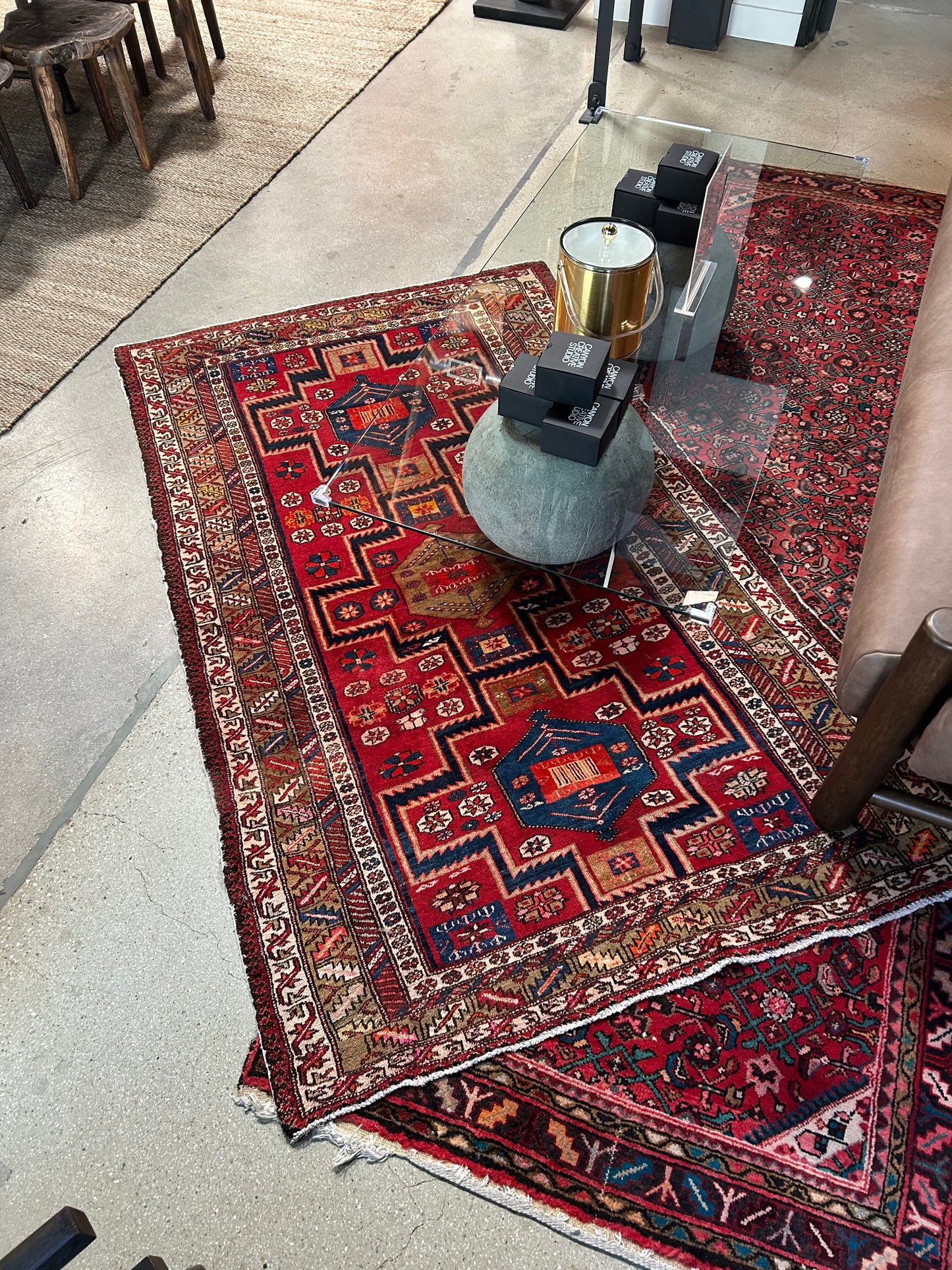A Vintage Aztec Rug (50x83) with bold geometric patterns is partially covered by a round gray table with black boxes, placed beside a brown leather sofa on a polished concrete floor in a modern interior.
