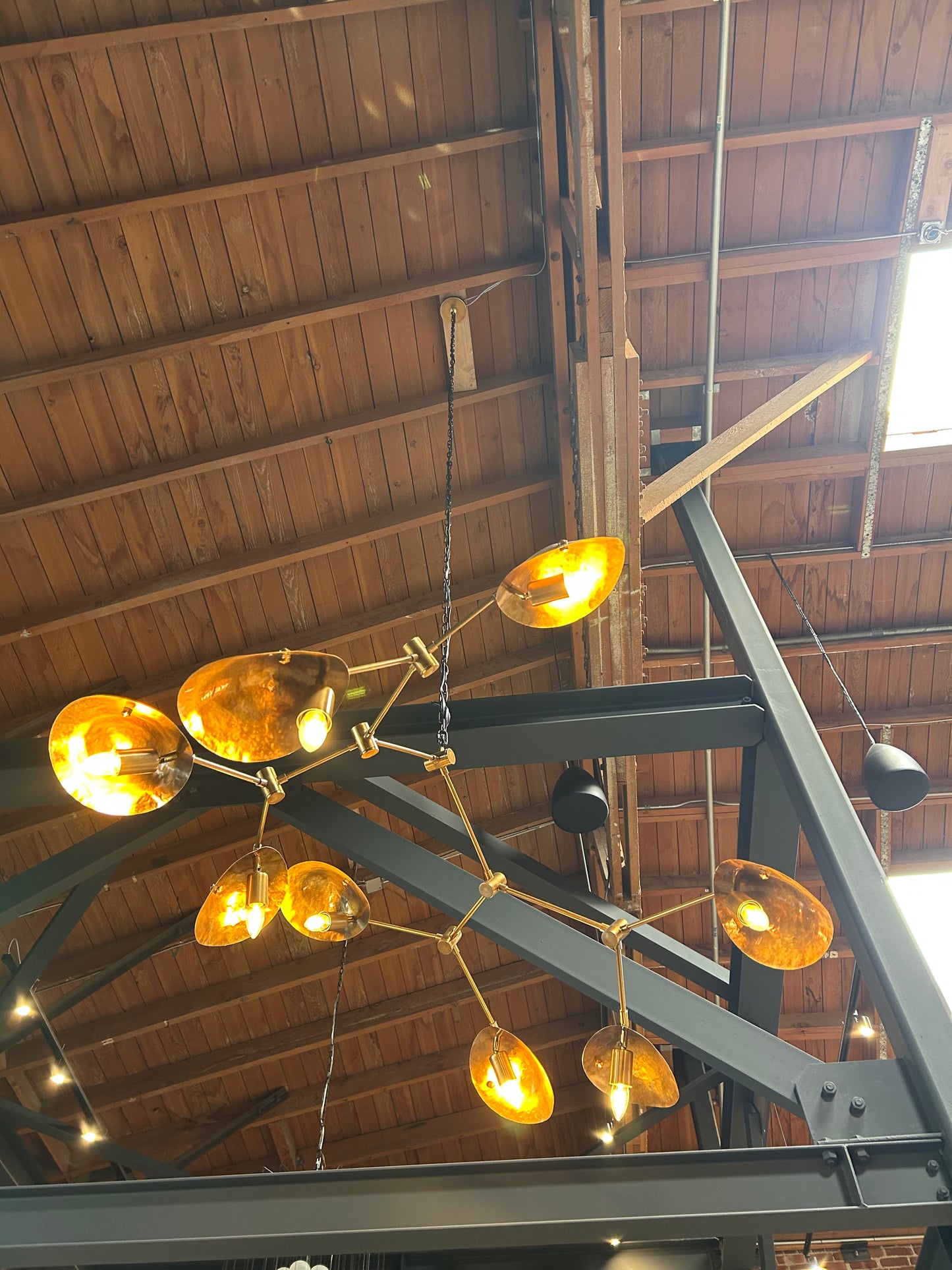 The 8-Arm Aged Brass Mantis Chandelier with amber glass shades hangs from a high ceiling with exposed wooden beams and black metal framework, casting warm light over the industrial-style space.