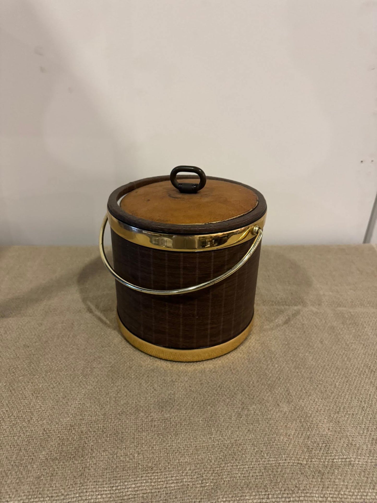 The Ice Bucket, 8.8 inches round in brown with a lid, gold accents, and a handle, rests on a beige textured surface against a plain white wall.