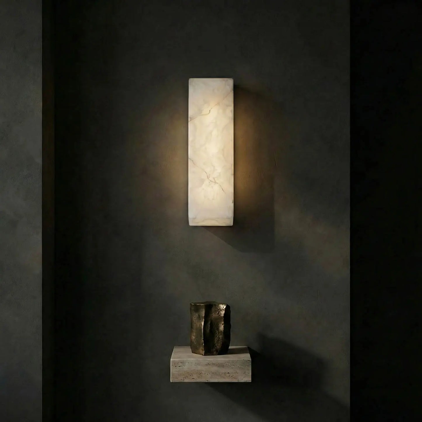 The Rectangular Sconce casts a soft, warm glow on a dark grey wall above a stone shelf with a textured metallic vase, its minimalist design enhancing the elegant simplicity of the space.