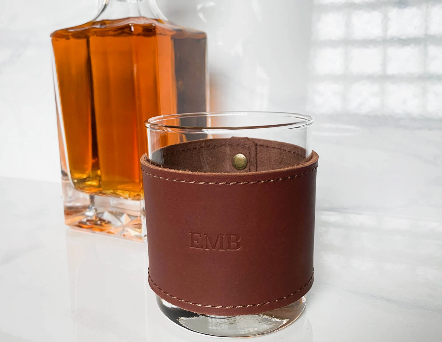 A glass of whiskey with a Leather Glass Cover embossed with EMB sits before a decanter of amber liquid on a white surface.