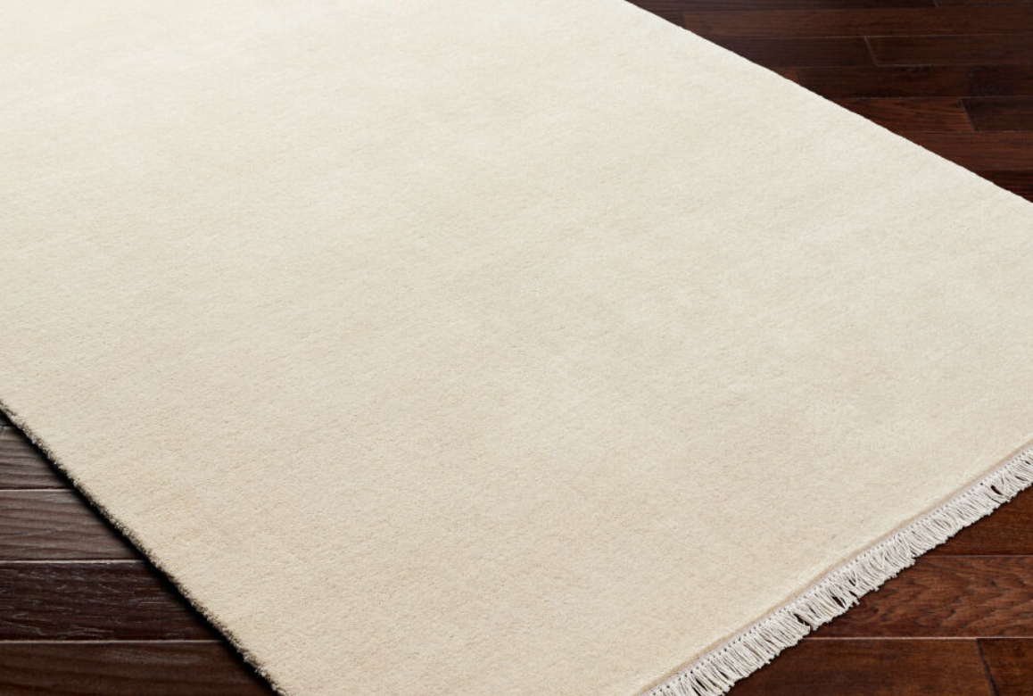 Fringe Ivory Wool Rug 9x12, a hand-knotted ivory wool rug with a fringed edge resting on a dark wood floor.