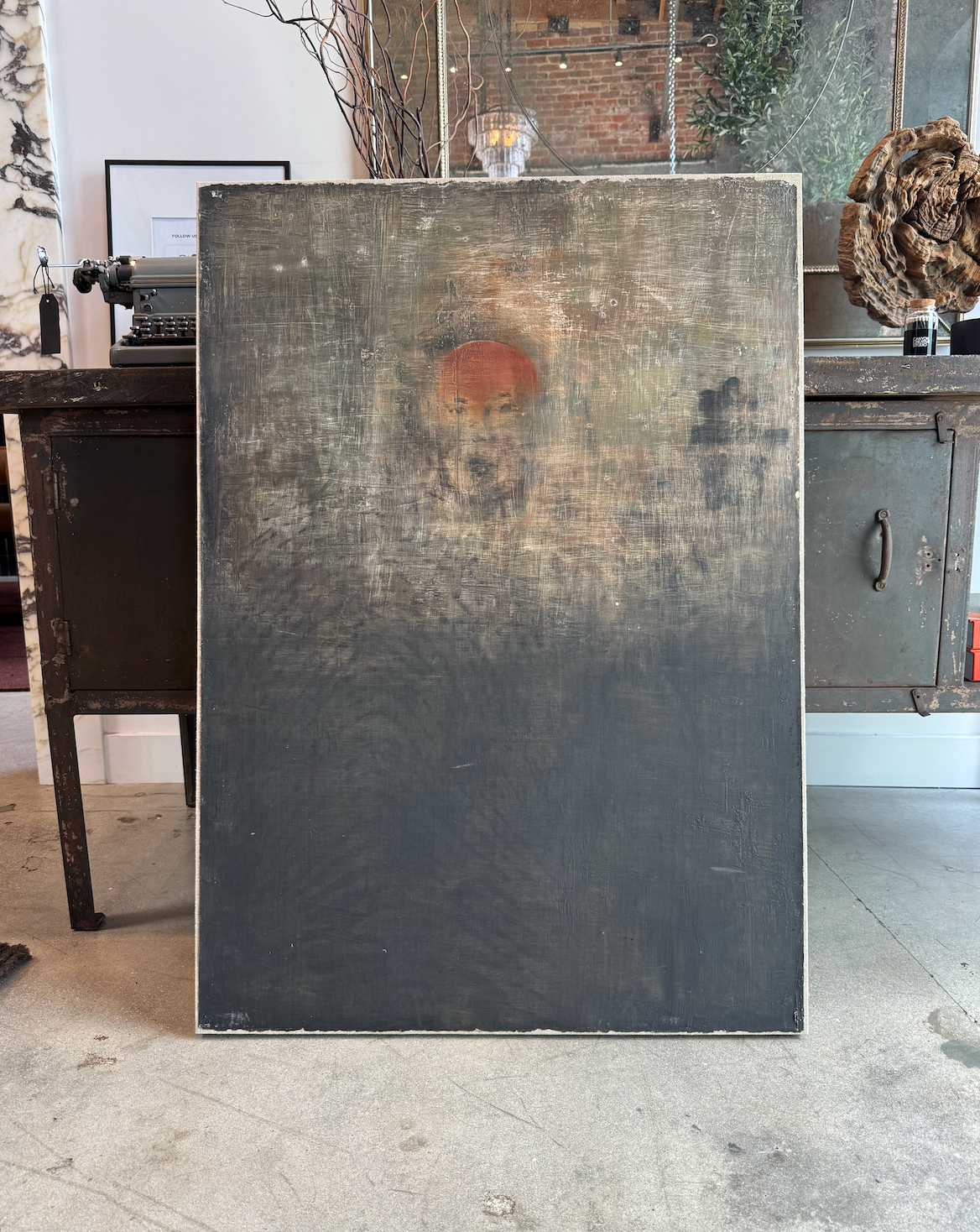 The Faded Face on Grey Ombre Canvas, measuring 43.00"h x 30.50"w, features a dark lower half and a faint abstract figure with a reddish head, displayed against an industrial metal cabinet in an eclectic room with exposed brick.