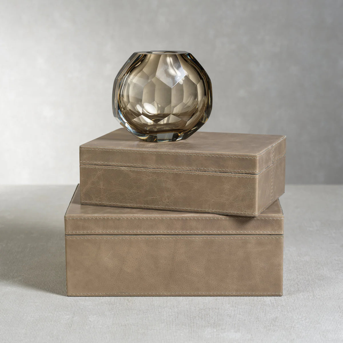 A large faceted glass vase with a smoky tint rests atop two stacked Mink Leather Boxes on a neutral surface against a light gray background, emphasizing the striking dimensions of the arrangement.
