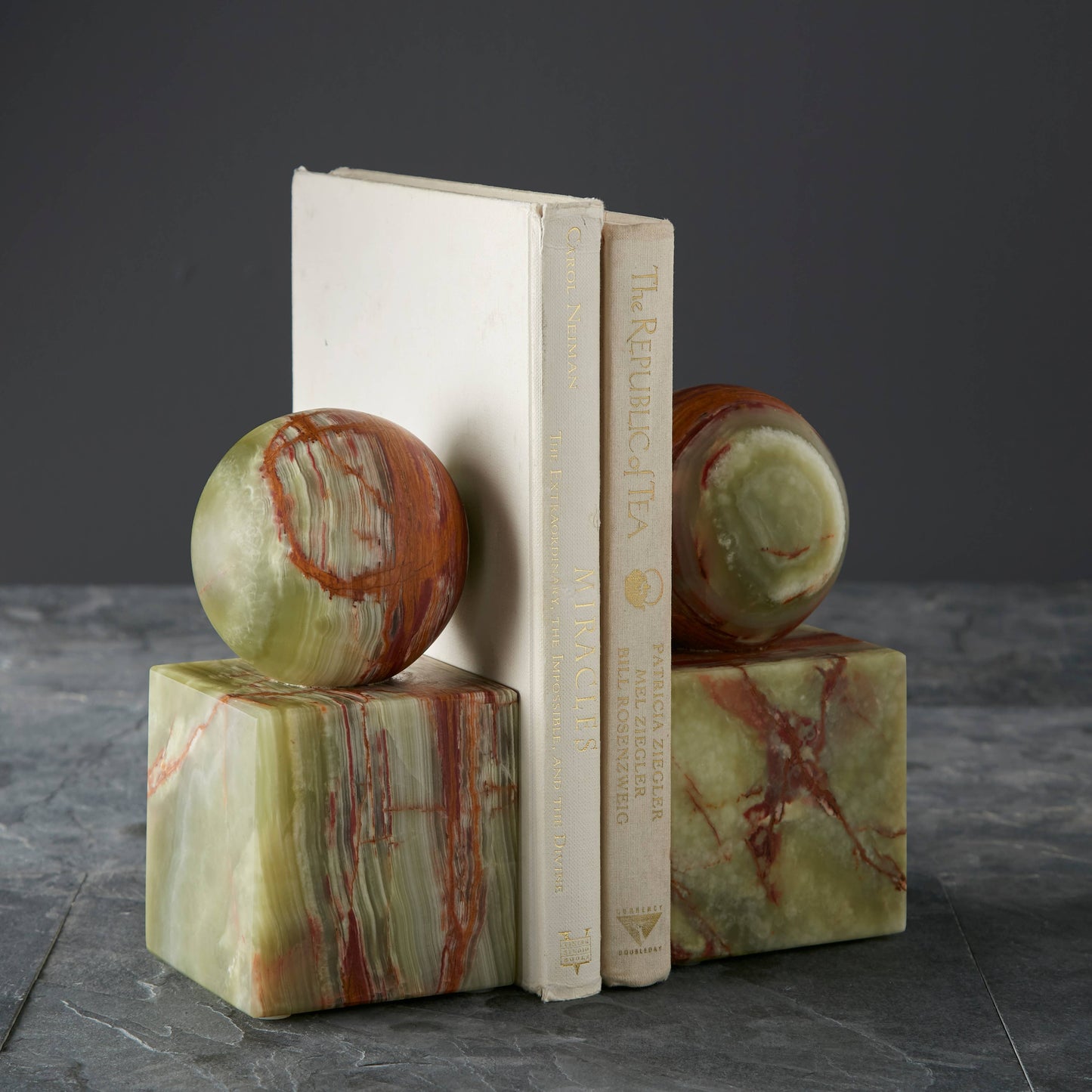 Two books stand upright between matching Verde Onyx Bookends—green cubes with red and cream veining in a honed finish, each topped by a sphere of the same stone, all displayed on a dark surface against a gray backdrop.