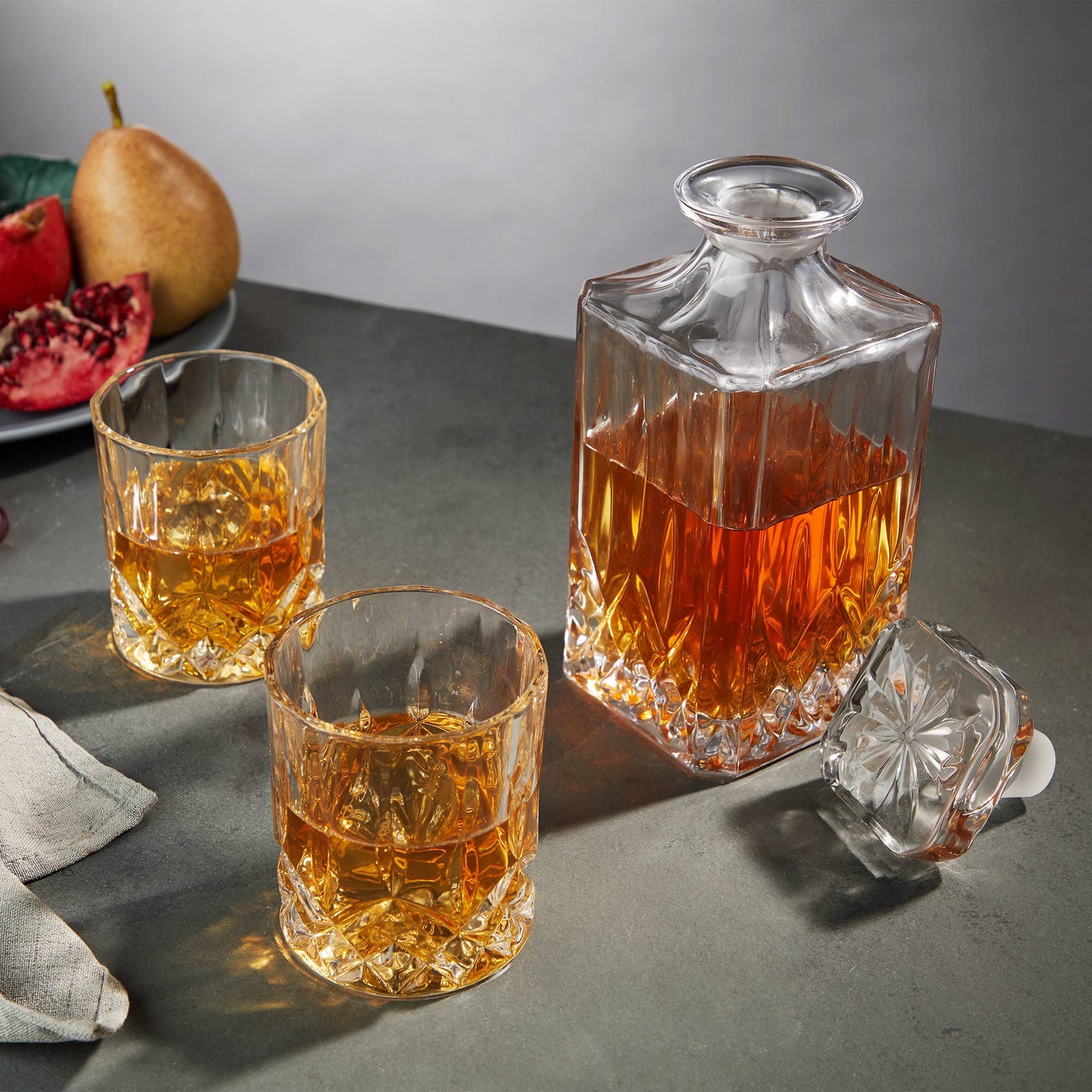 The Crystal Whiskey Decanter and Tumblers (Set of 3), filled with whiskey, rests on a gray surface beside its lid and two matching glasses. Behind it is a plate of fruit featuring a pear and a pomegranate.