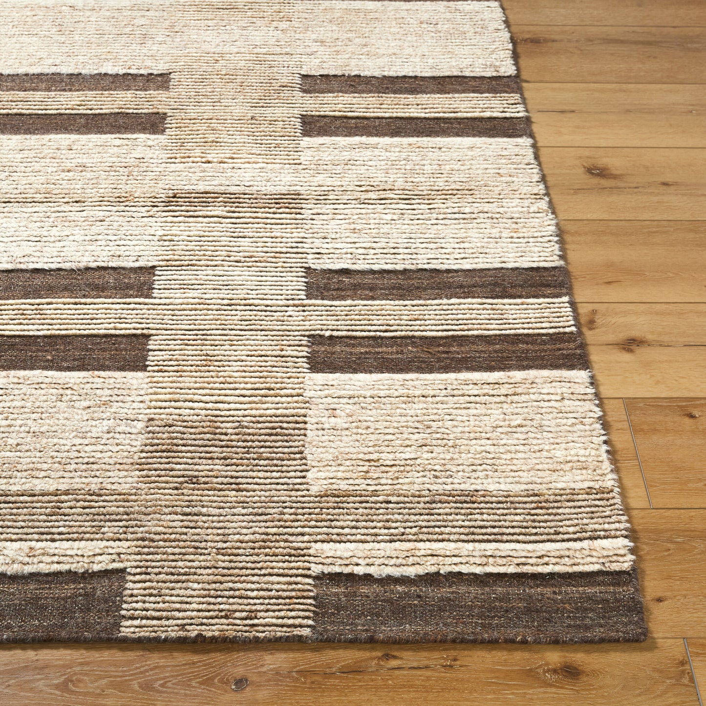 The Patched Rug 8 x 10, a beige and brown hand-knotted wool piece with alternating geometric stripes and rectangles, sits on a light wood floor.