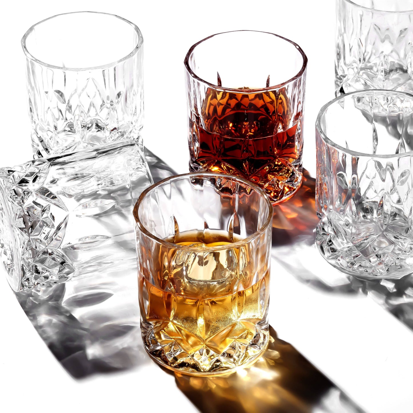 A Crystal Cut Old Fashioned Whiskey Glass 10oz (1pc) on a white surface—three upright (one empty, one with amber liquid, one with dark brown liquid) and two lying on their sides, casting detailed shadows. Ideal for whiskey lovers.