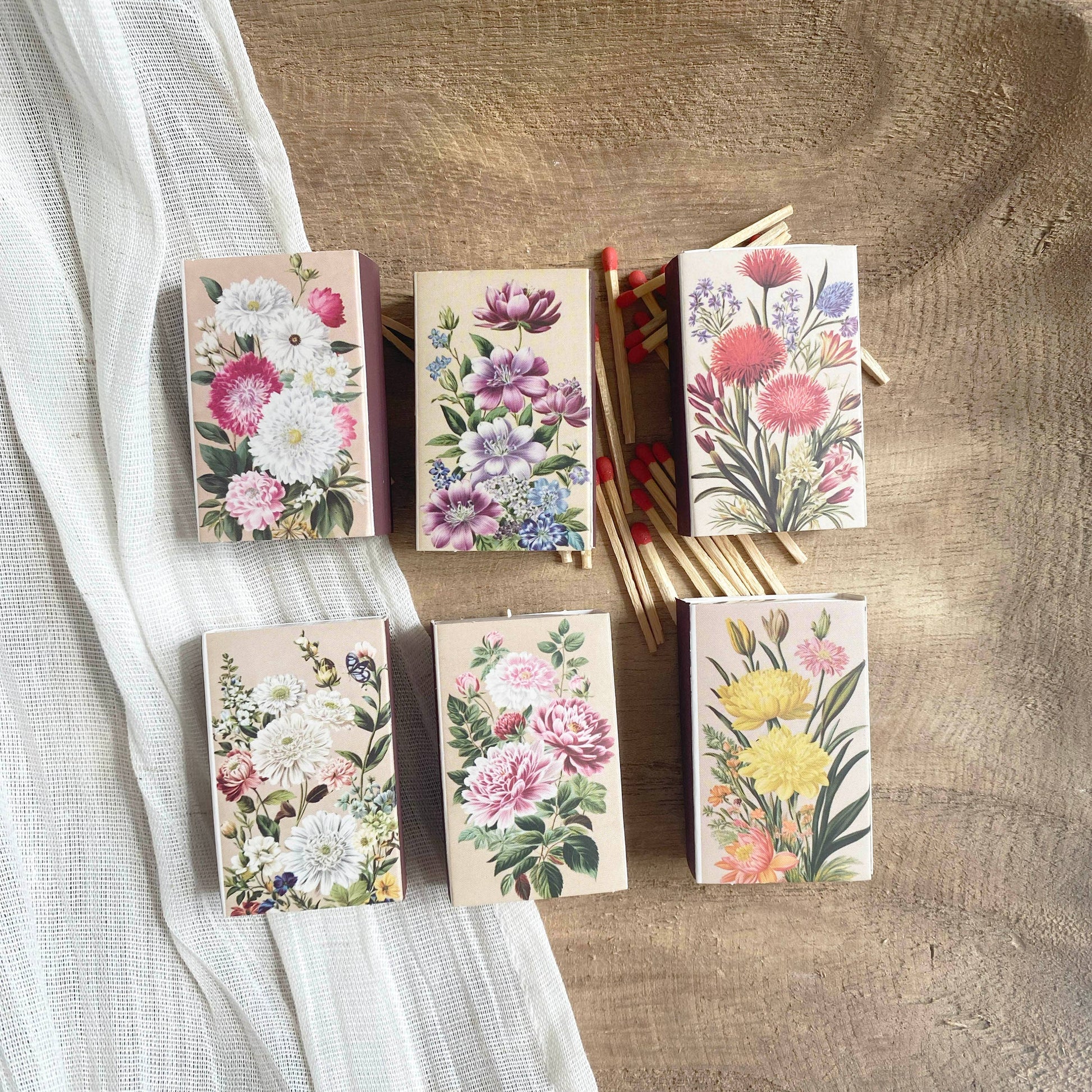Six Vintage Matchbook sets featuring botanical flower illustrations are displayed on a wooden surface. Red-tipped strike-on-box matches are visible, with a white textured fabric draped on the left. Perfect for floral design lovers.