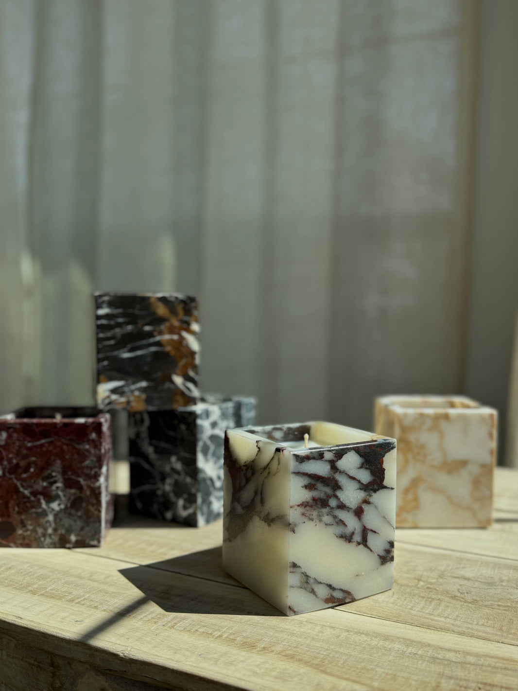 Marble Candles, available in white, black, and brown, double as elegant reusable vessels. Displayed on a light wooden surface amid soft sunlight and sheer curtains, they add a sophisticated decorative touch to any space.