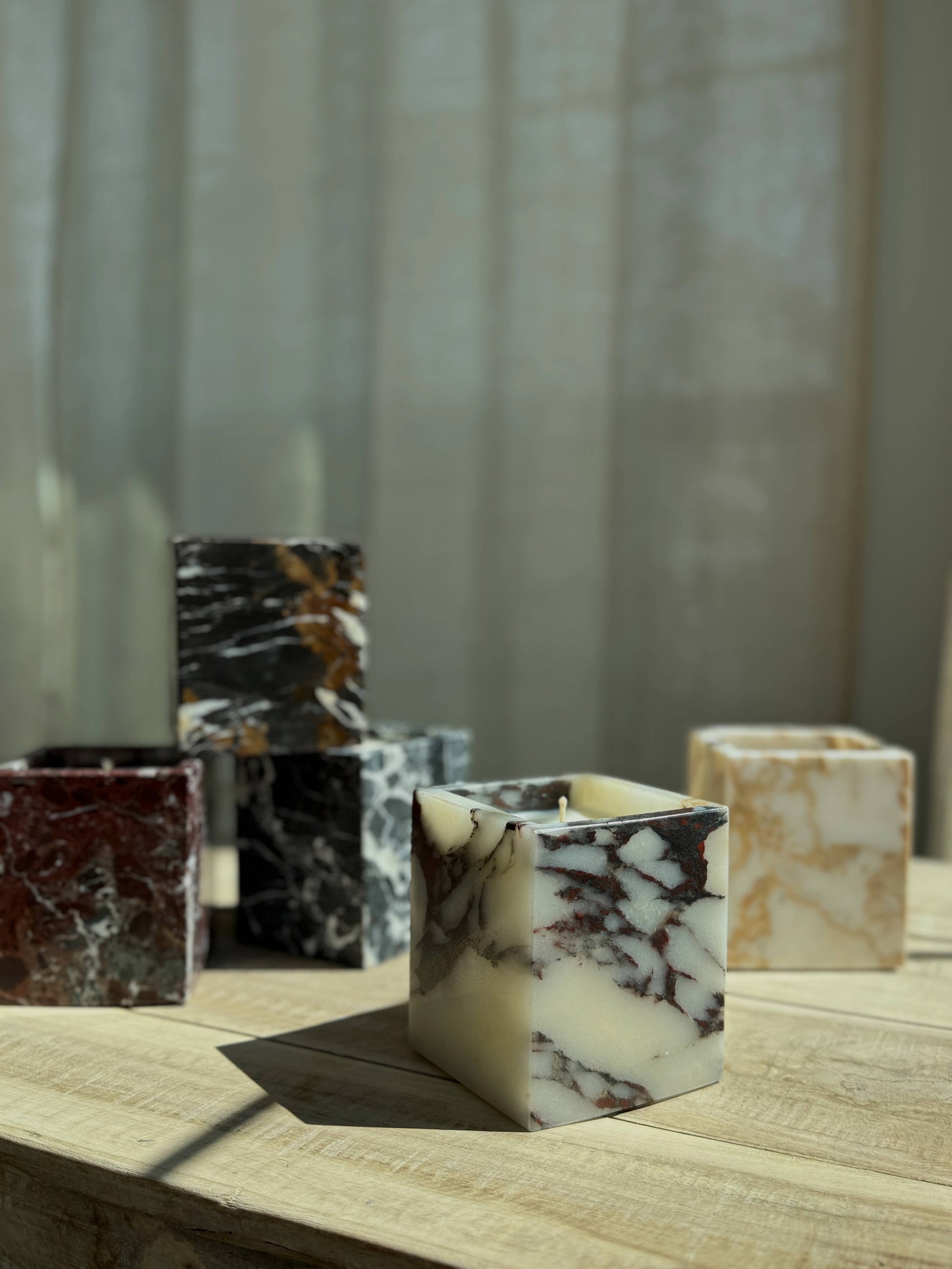 Marble Candles, available in white, black, and brown, double as elegant reusable vessels. Displayed on a light wooden surface amid soft sunlight and sheer curtains, they add a sophisticated decorative touch to any space.
