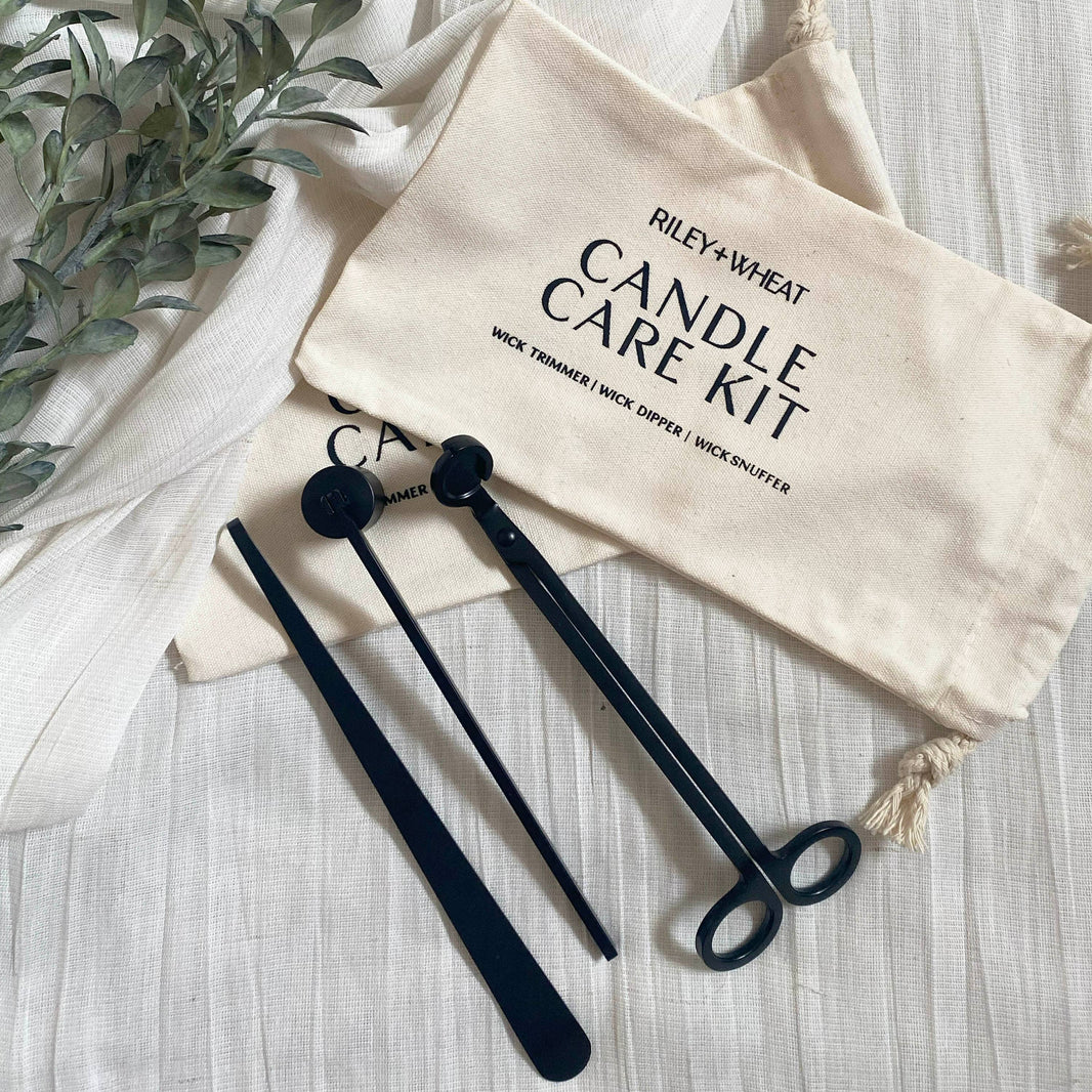 The Candle Care Kit includes a black metal wick trimmer, dipper, and snuffer, resting on a cream cloth pouch labeled "Candle Care Kit," with green leaves beside it.