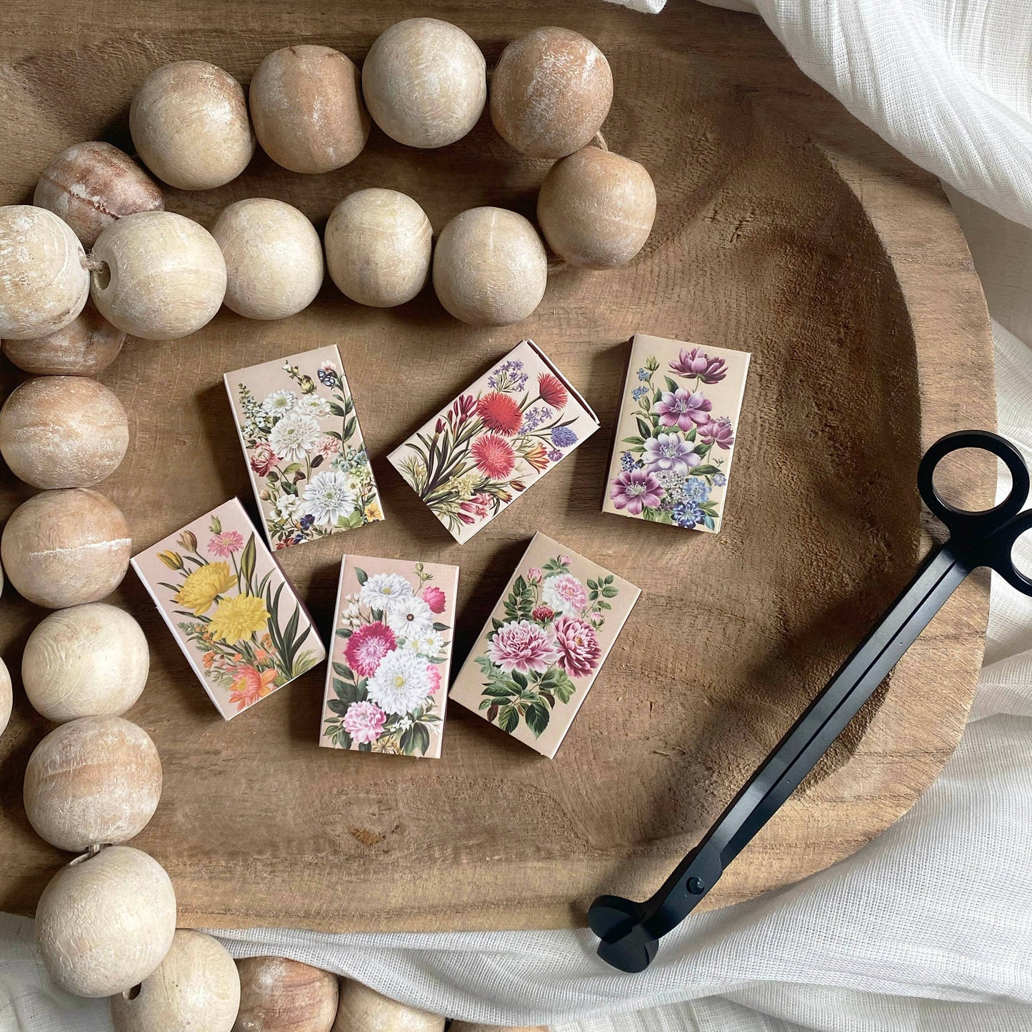 Six Vintage Matchbook sets in matchboxes are arranged on a wooden tray with large beads and black candle wick trimmers, all atop white textured fabric for a charming vintage-inspired display.