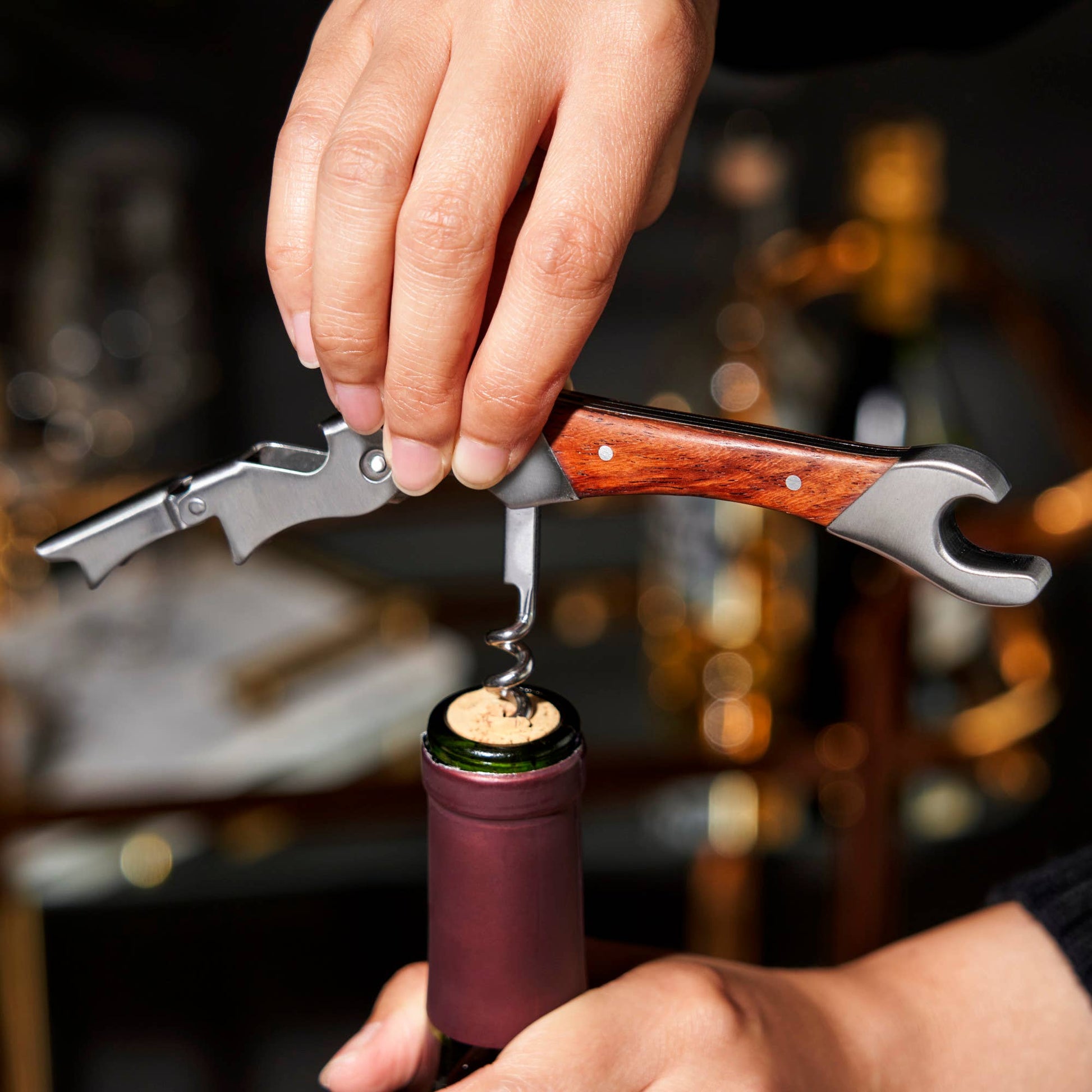 Using the Rosewood DoubleHinged Corkscrew and Foil Cutter, a person steadies a wine bottle with one hand while twisting the tool’s wooden handle into the cork with the other to open it.