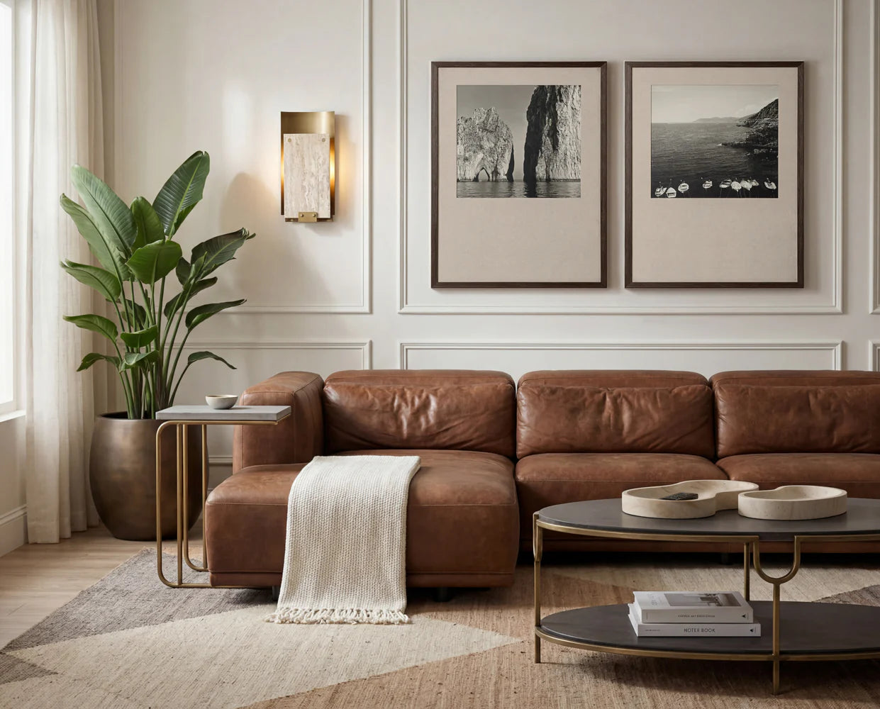 A stylish living room with a brown leather sofa, beige throw, modern tables, potted plant, Travertine Sconce on a white paneled wall, and two framed nature photos. Natural light filters in from the left.
