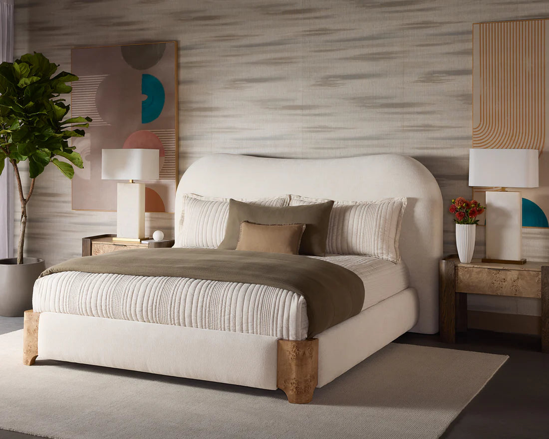 A modern bedroom with a curved white bed, striped bedding, olive green blanket, neutral throw pillows, and a pair of Two-Toned Wooden Nightstands with lamps. The decor includes abstract wall art, a leafy plant, and a gray textured wall.