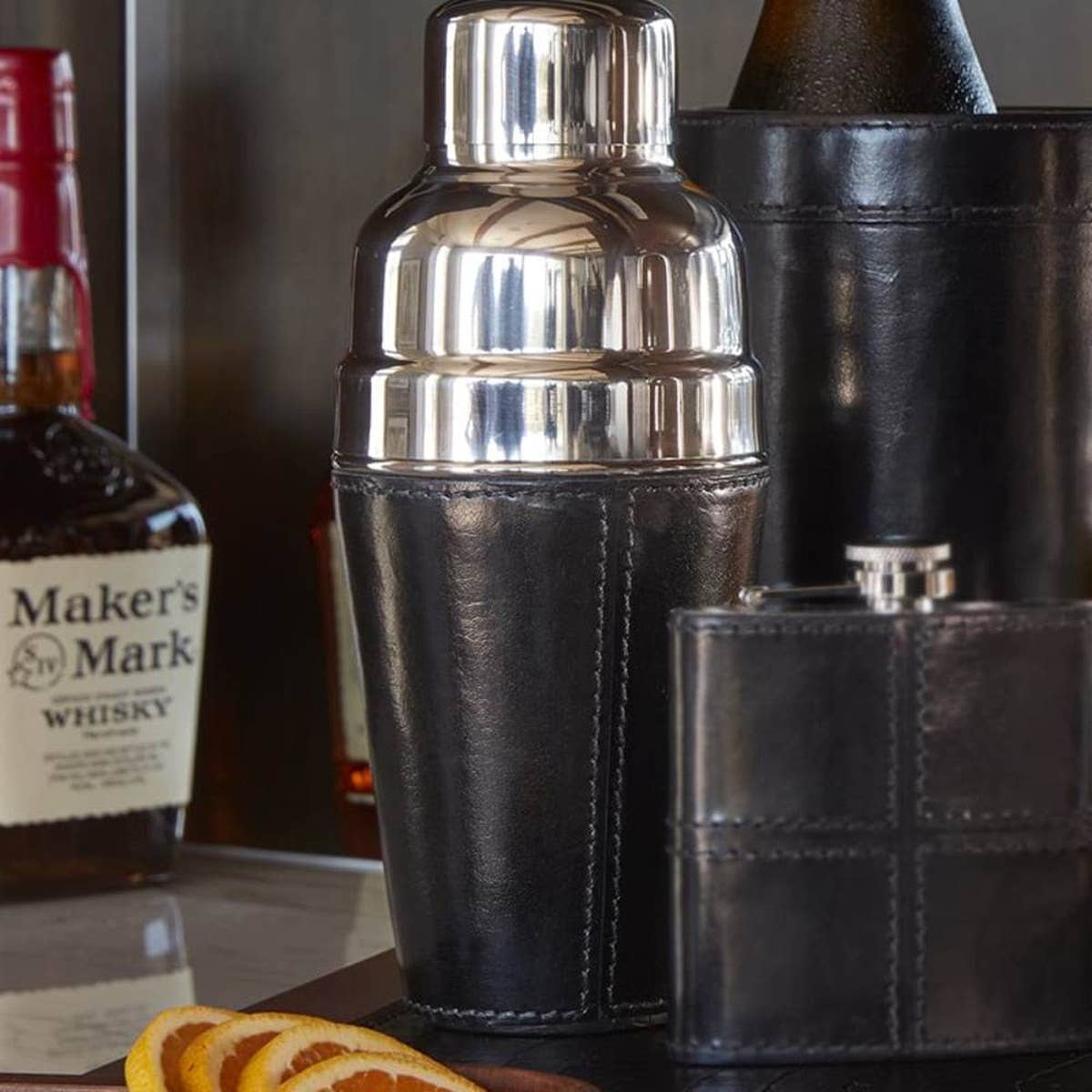 The Leather Cocktail Shaker Black sits on the counter alongside a matching leather flask, a bottle of Maker’s Mark whisky, and orange slices—making it an ideal bar gift for any cocktail enthusiast.