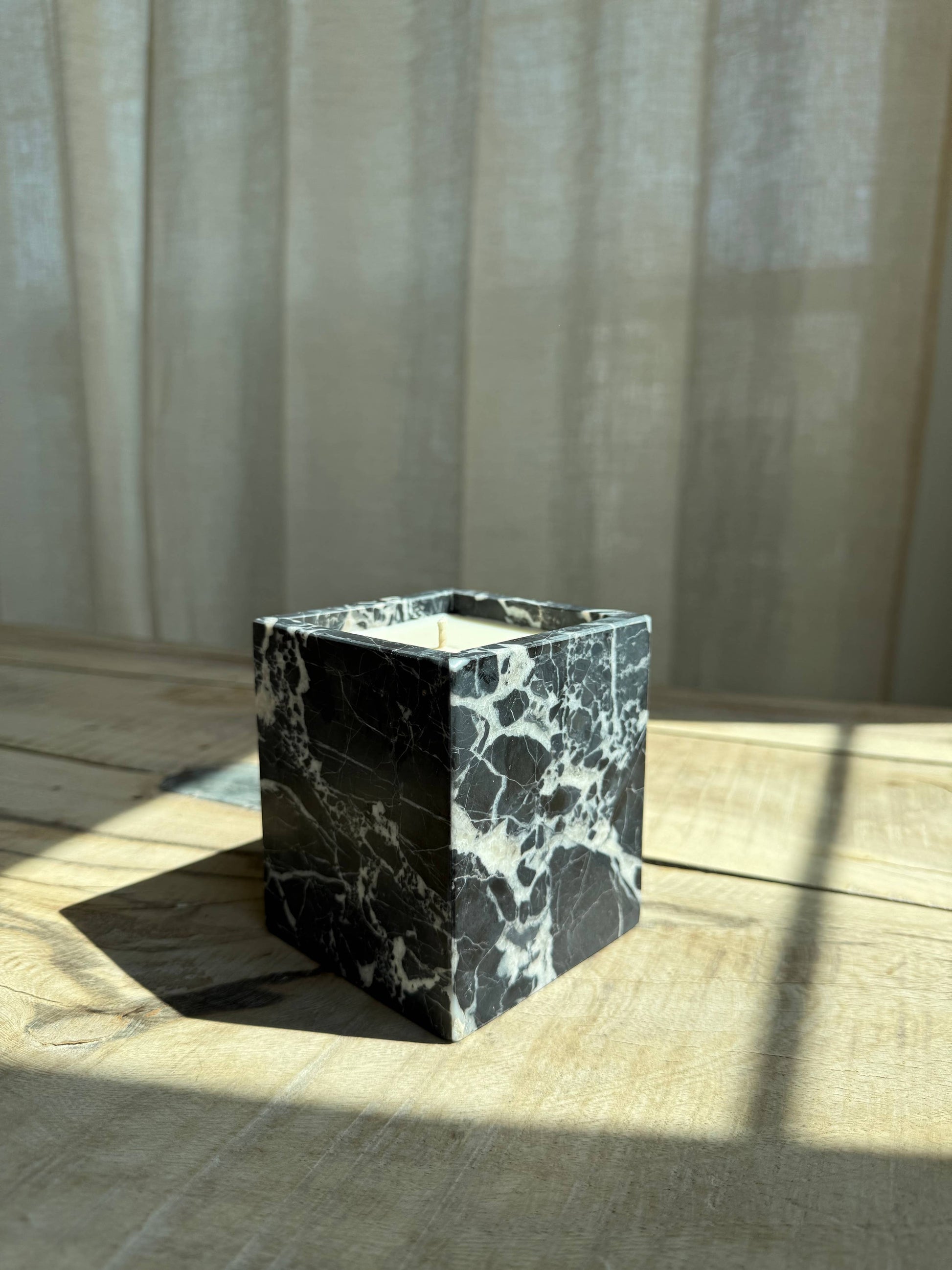 The Marble Candles, a small square vessel with white veins, serve as elegant candle holders, displayed on wood and beautifully lit by sunlight filtering through a sheer curtain in the background.