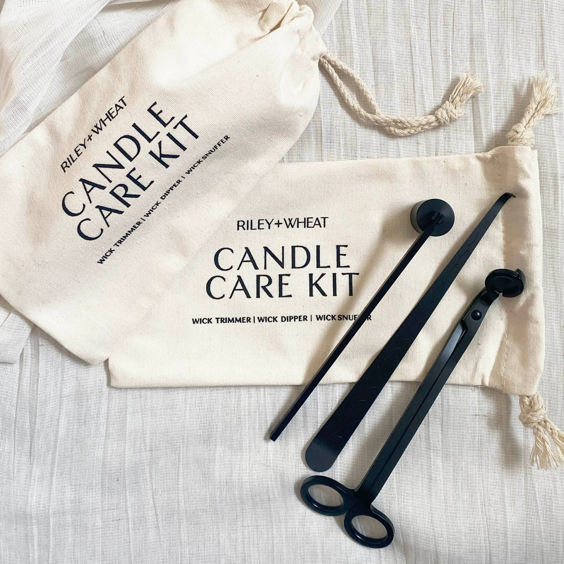 The Candle Care Kit includes a sleek black wick trimmer, dipper, and snuffer on a white cloth, alongside two cream Riley + Wheat Candle Care Kit pouches.