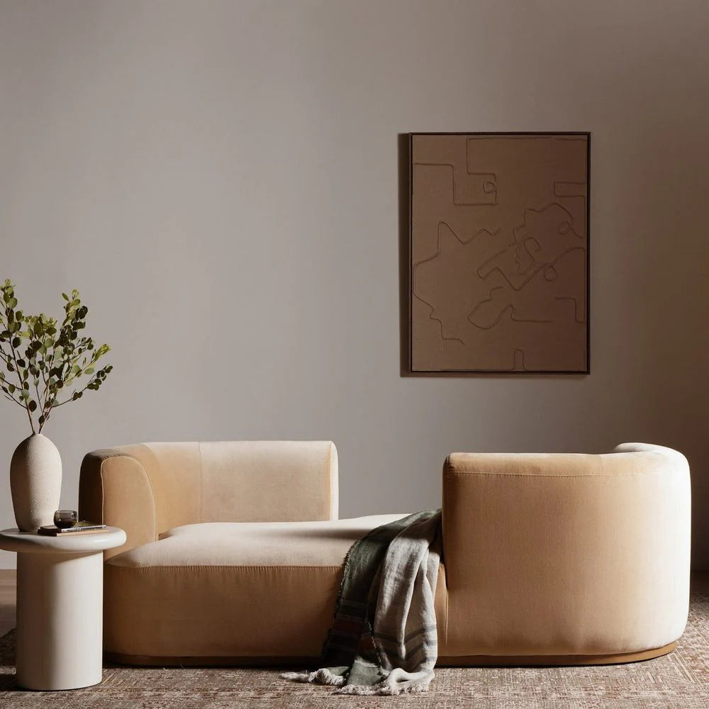 The Deandra Tete A Tete Chaise in beige (90.5") sits on a textured rug with a gray throw draped over one side. Next to it is a round side table with a white vase of green branches, while abstract art decorates the neutral wall behind.