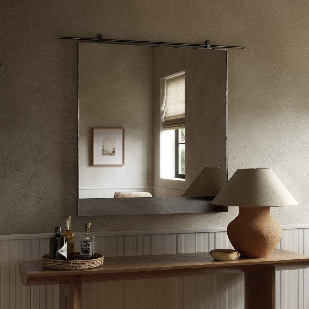 The Chico Mirror hangs above a wooden console table holding a beige lamp, woven tray, and bottles. The mirror reflects a window with a shade and framed picture on the light-colored wall.