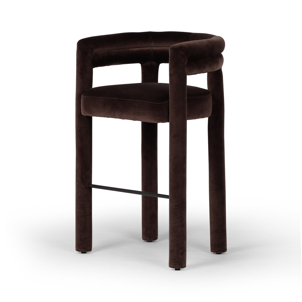 The Upholstered Bar Stool features a minimalist, contemporary design with a rounded backrest and armrests, dark brown velvet upholstery, four thick cylindrical legs, and a metal footrest.
