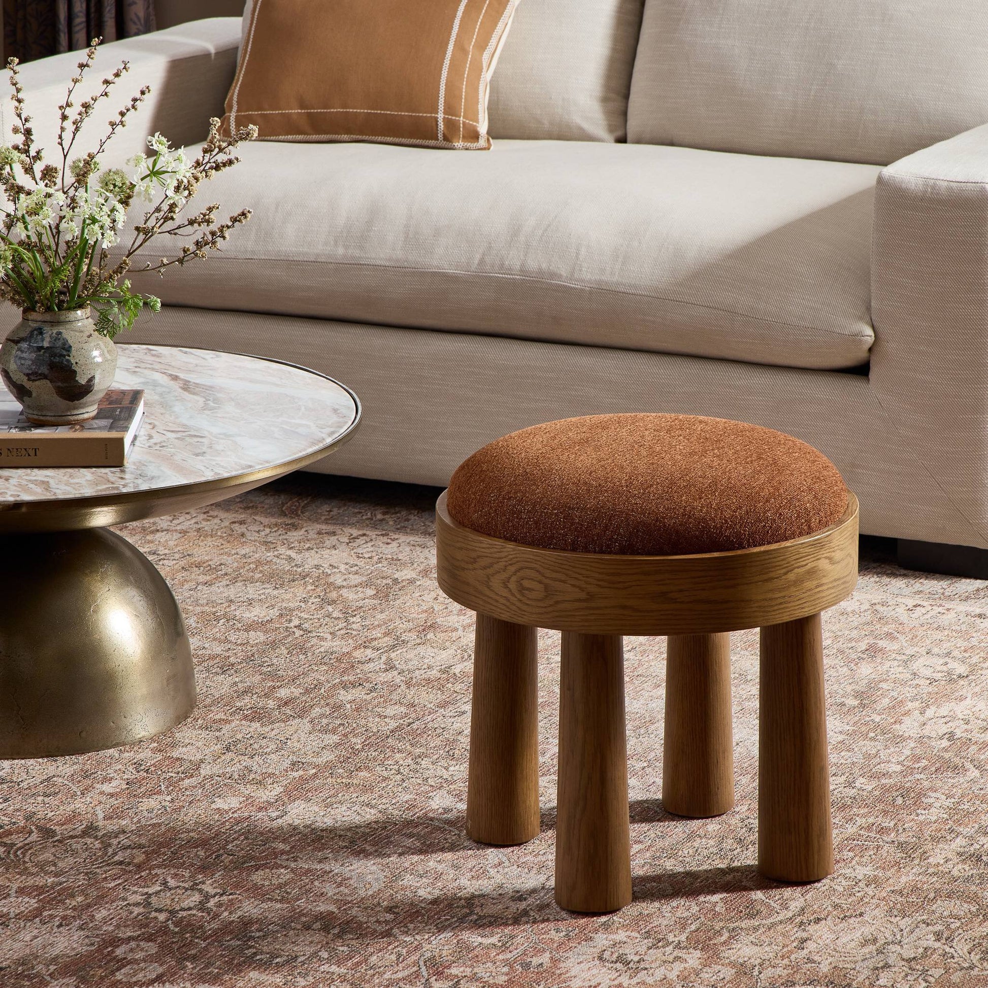 A Louise Accent Stool with a round wooden frame and brown cushioned seat sits on a patterned rug next to an accent table and marble coffee table with a vase, in front of a beige sofa with a matching pillow.