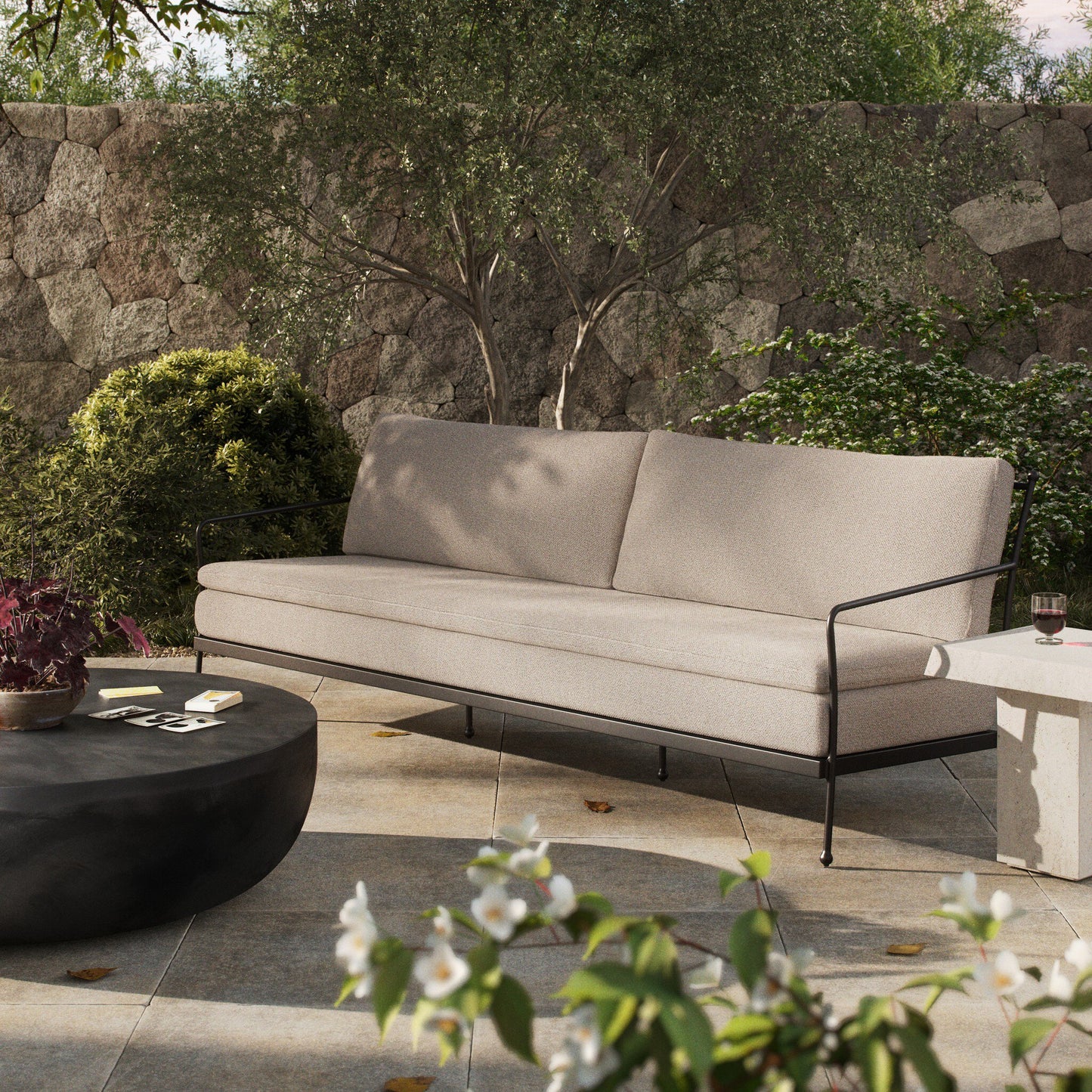 The Tally Outdoor Sofa, featuring beige upholstery and black metal arms, sits on a stone patio beside a round black coffee table, its sleek design complementing the bench and lush garden with flowering plants and a stone wall.