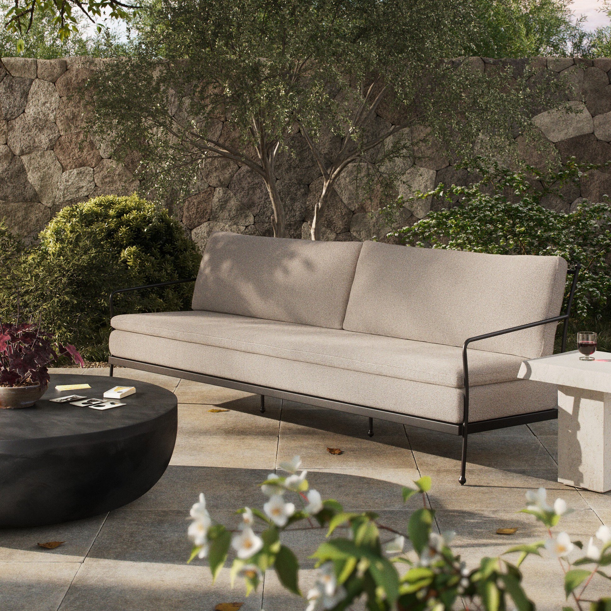 The Tally Outdoor Sofa, featuring beige upholstery and black metal arms, sits on a stone patio beside a round black coffee table, its sleek design complementing the bench and lush garden with flowering plants and a stone wall.
