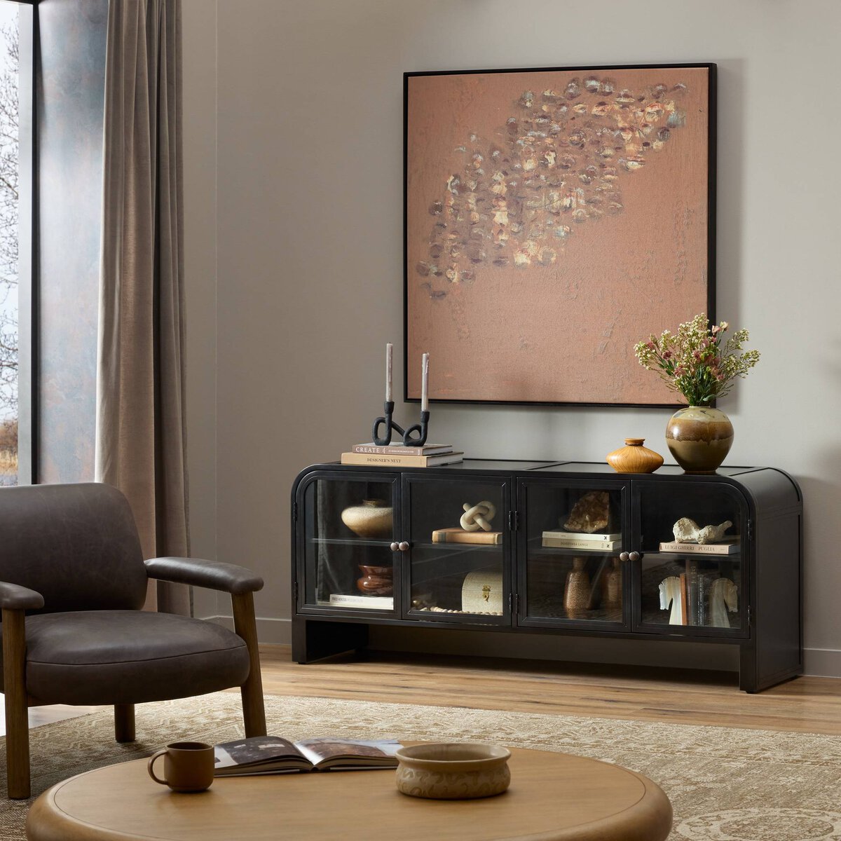 A modern living room featuring a Black Iron Media Console with glass doors, abstract wall art, candles, a floral vase, a wooden coffee table, and a dark armchair on a beige rug by the window with beige curtains.