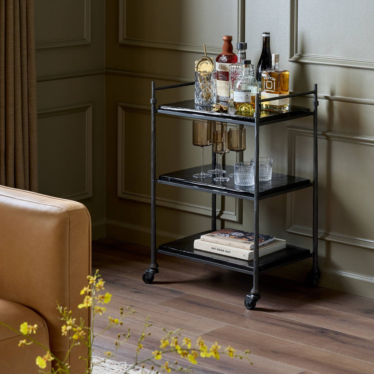 Teagan Bar Cart: a three-tiered black rolling bar cart, 23.00″w x 15.50″d x 33.00″h, holding glass bottles, glassware, and a book on the bottom shelf.