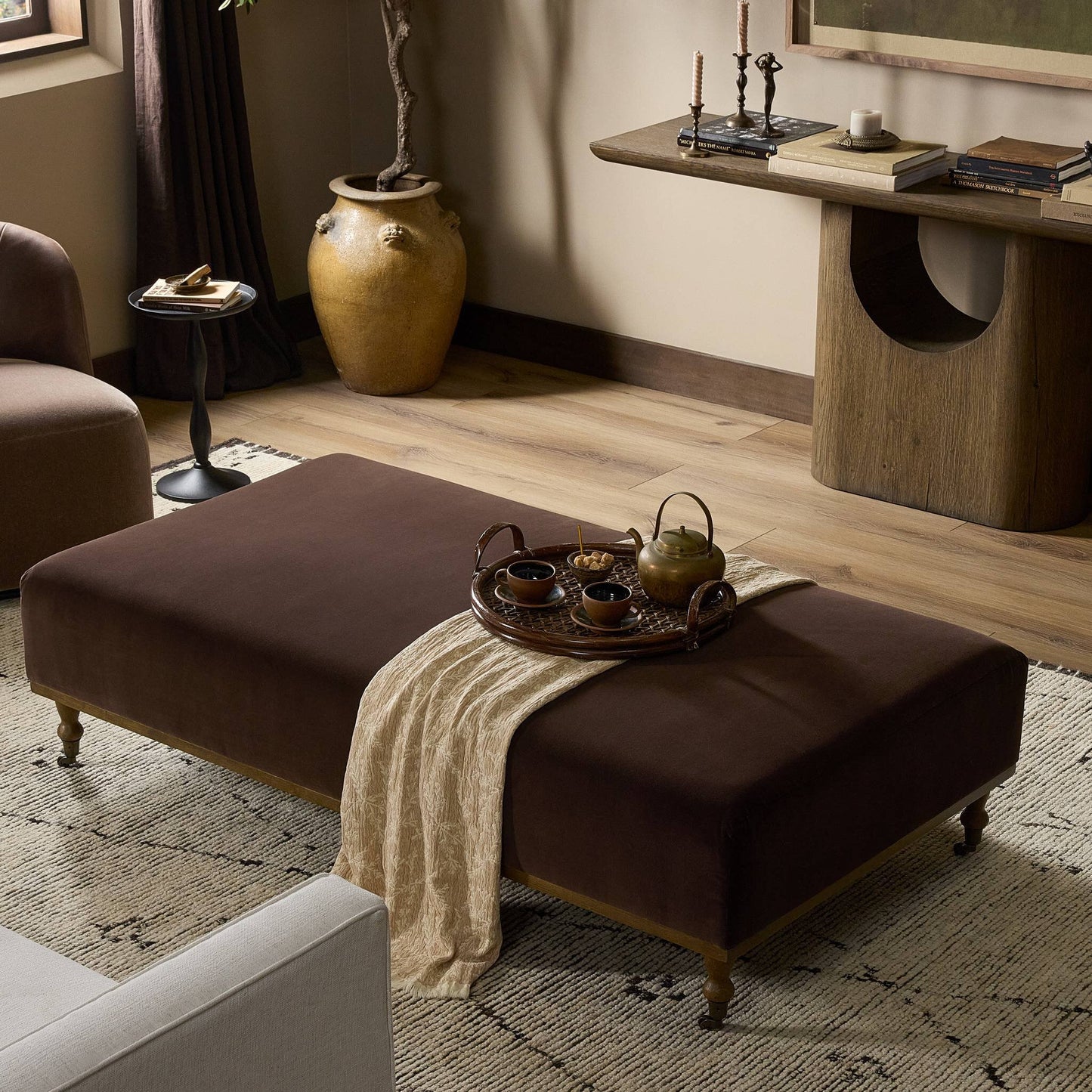 The Harlin Cocktail Ottoman, measuring 65.00"w x 35.00"d x 17.50"h with brown upholstery and wooden legs, serves as a coffee table topped with a tray holding a teapot and cups in a warmly decorated living room.